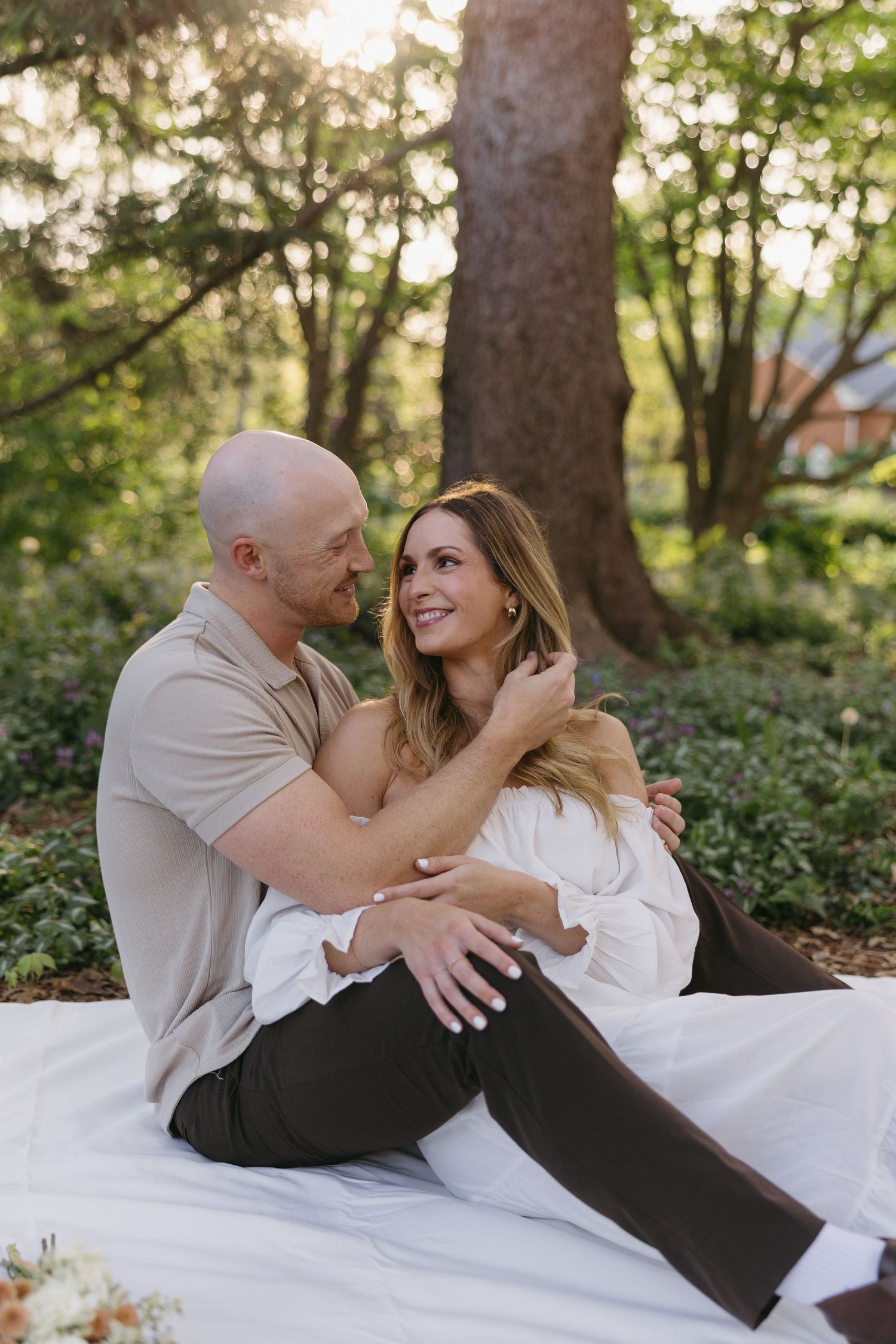 Couple sitting closely on a white blanket in a sunlit park, smiling and embracing each other warmly.