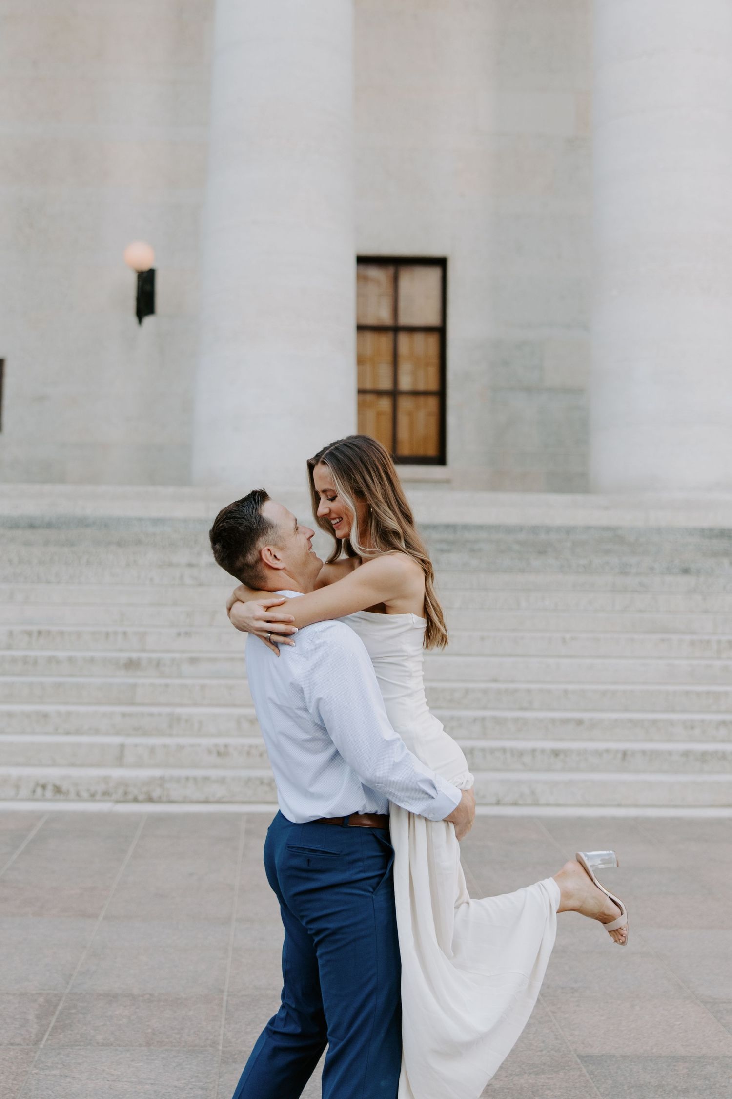 Groom lifting bride in white dress while she laughs joyfully on steps of a marble building.