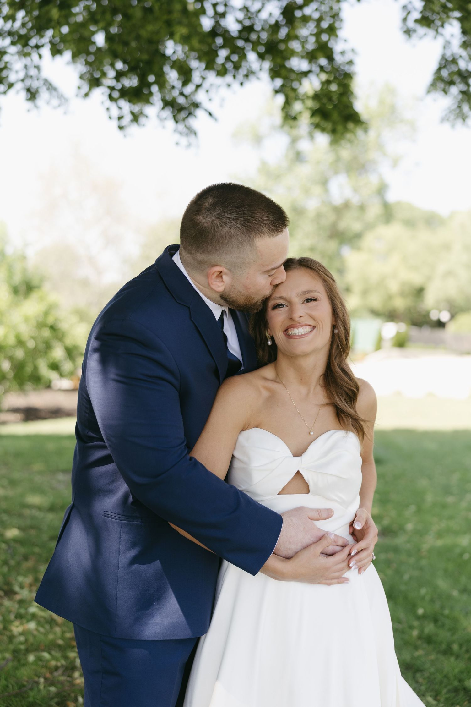 Groom in navy suit embraces and kisses bride in a strapless cutout gown under a green tree.
