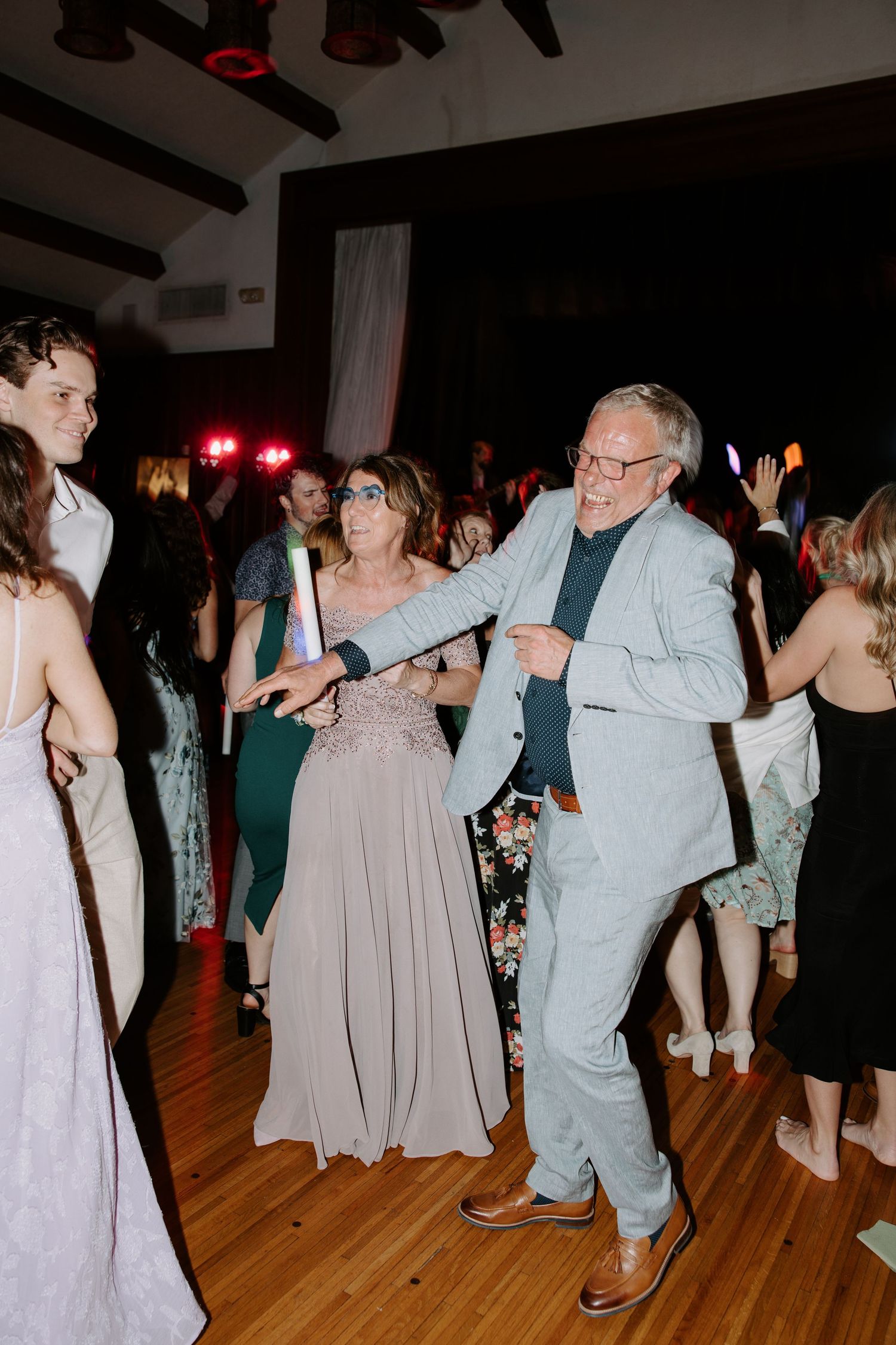 Couple dancing joyfully at a wedding reception on a wooden dance floor surrounded by guests in formal attire.