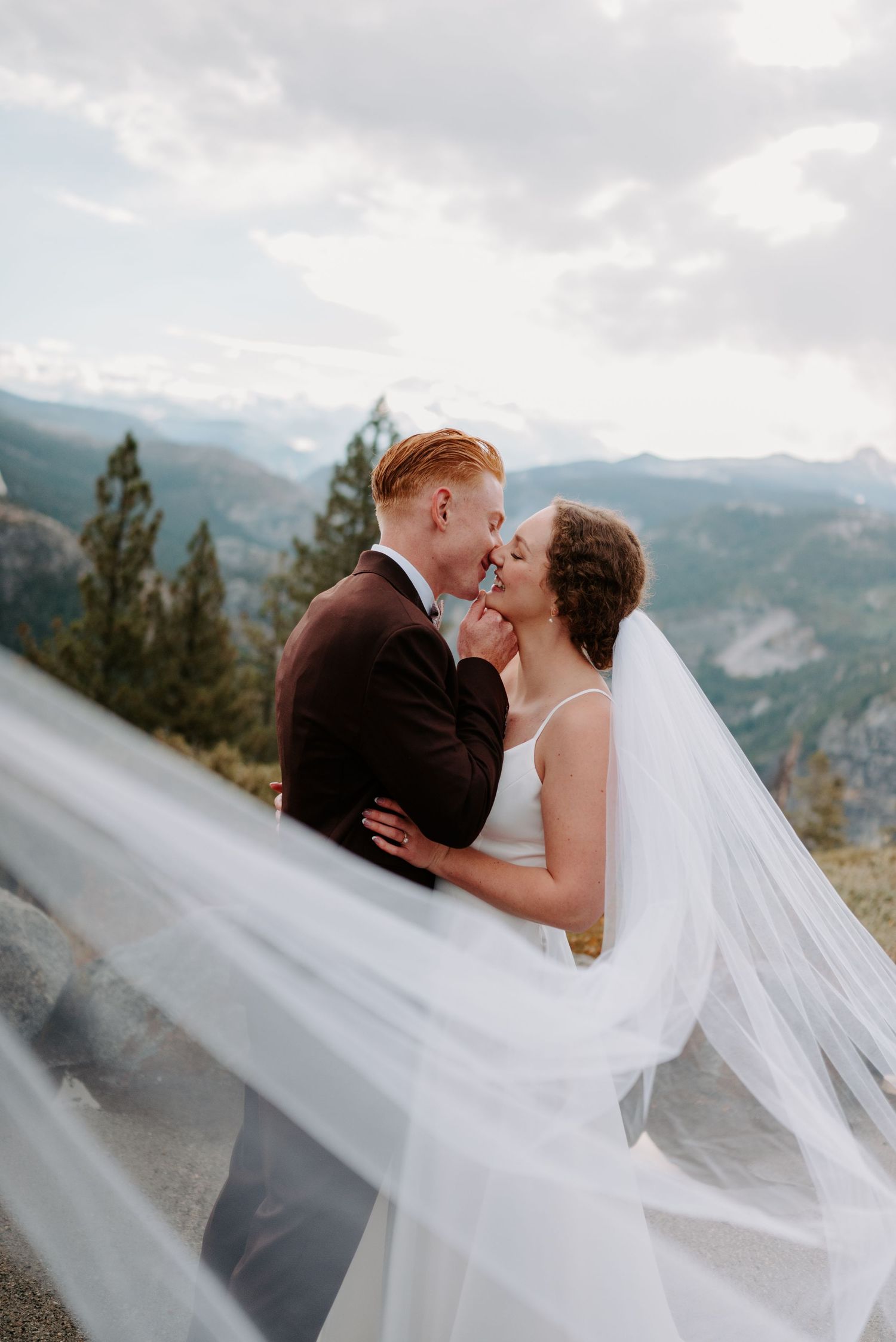 Bride and groom share a kiss with flowing veil and mountain landscape at outdoor wedding.