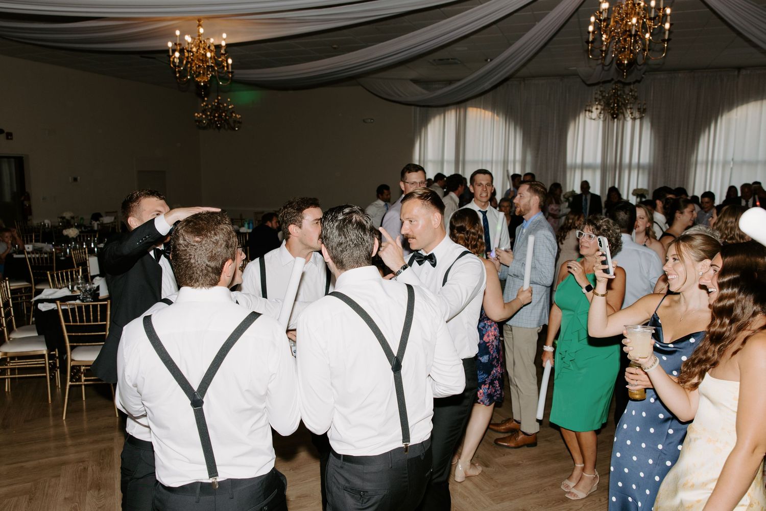 Wedding guests in formal attire dancing and celebrating on the dance floor at a bright elegant reception venue.