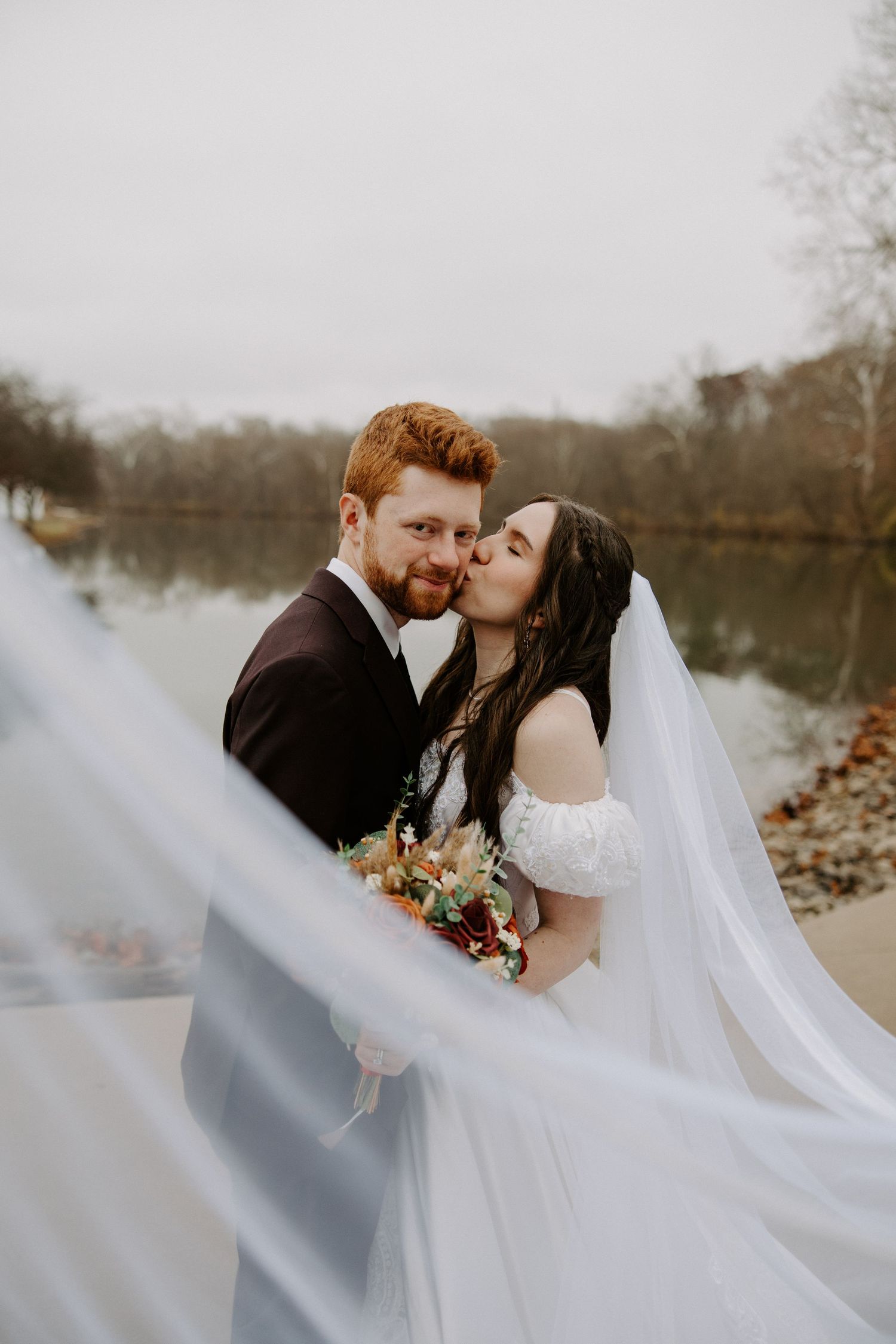 Groom with red beard kisses bride in white off-shoulder gown near lake as veil billows in the foreground.