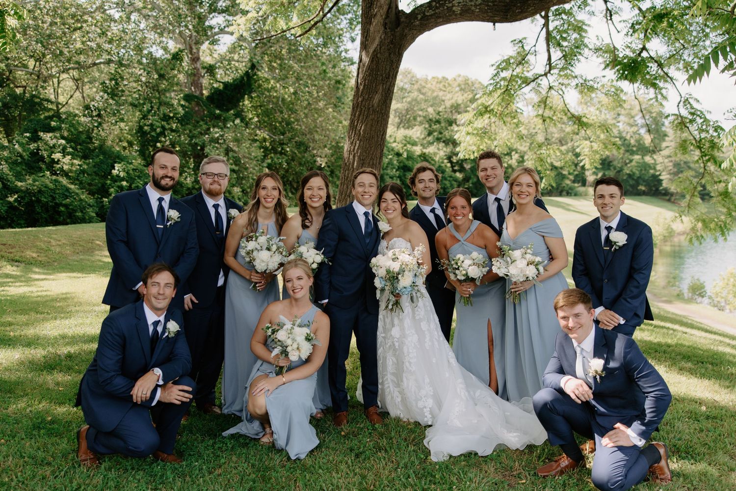 Wedding party poses under a tree outdoors, bride in lace gown with groomsmen in navy suits and bridesmaids in dusty blue dresses.
