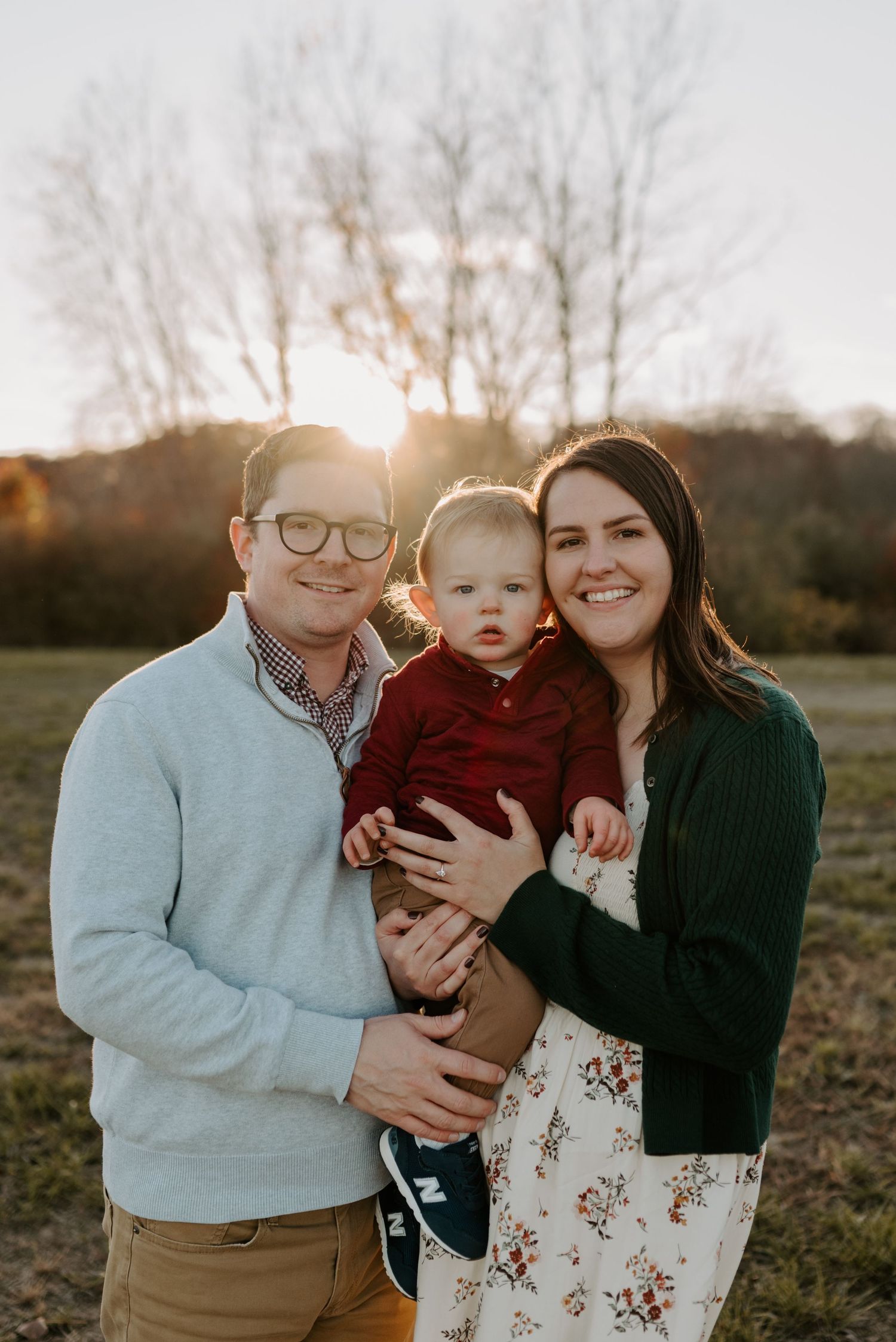 A young family with a baby boy poses outdoors at golden hour in a field with bare trees in the background.