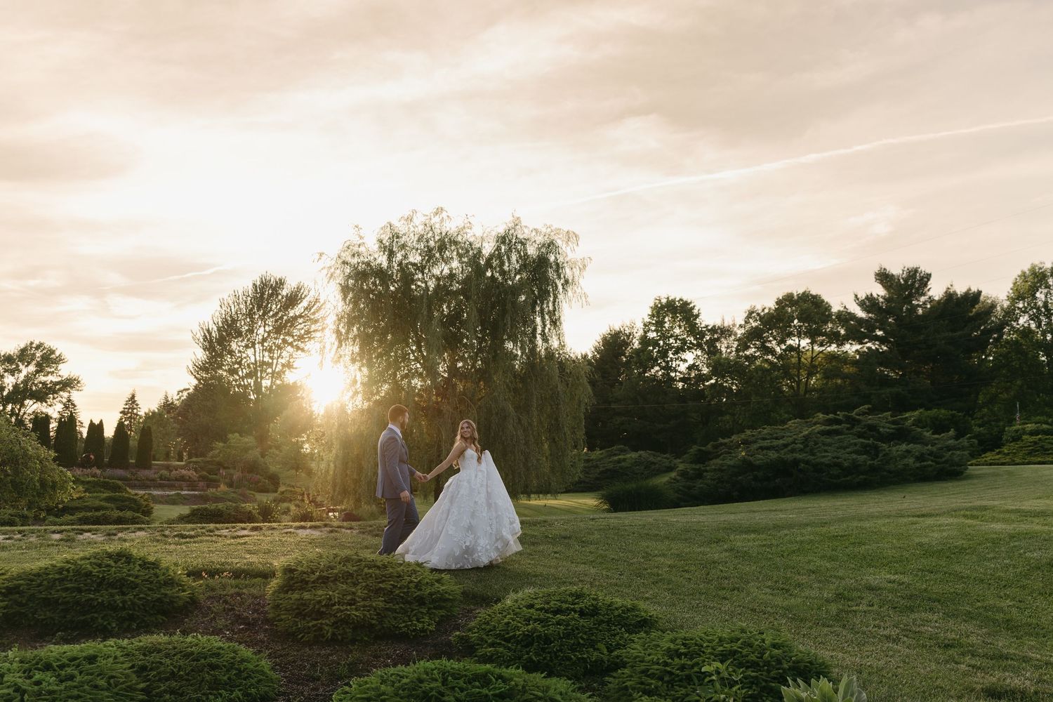 Bride and groom walk hand in hand through lush garden at golden sunset on their wedding day.
