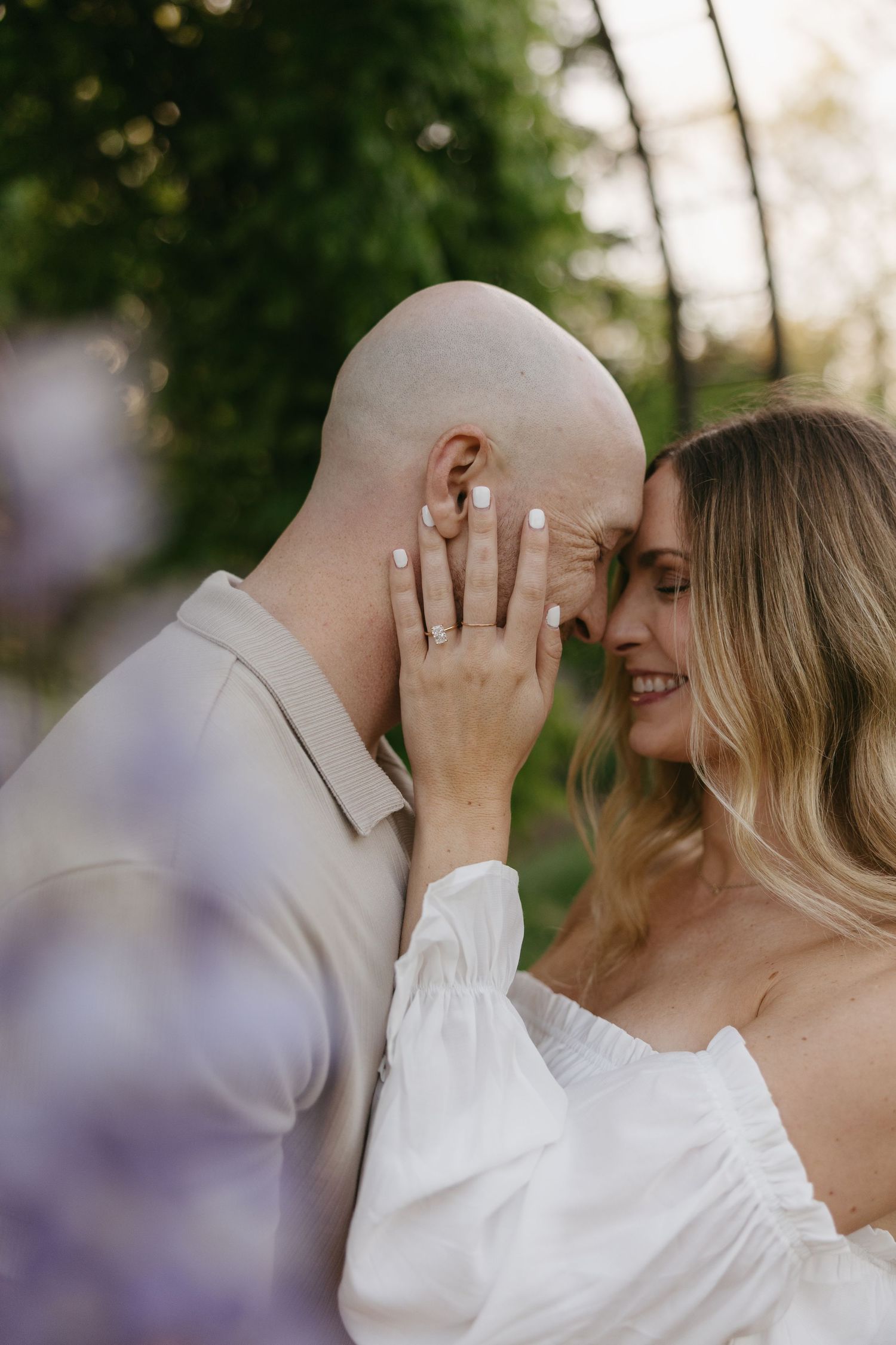 Woman showing off engagement ring while embracing bald man outdoors surrounded by soft purple blooms.
