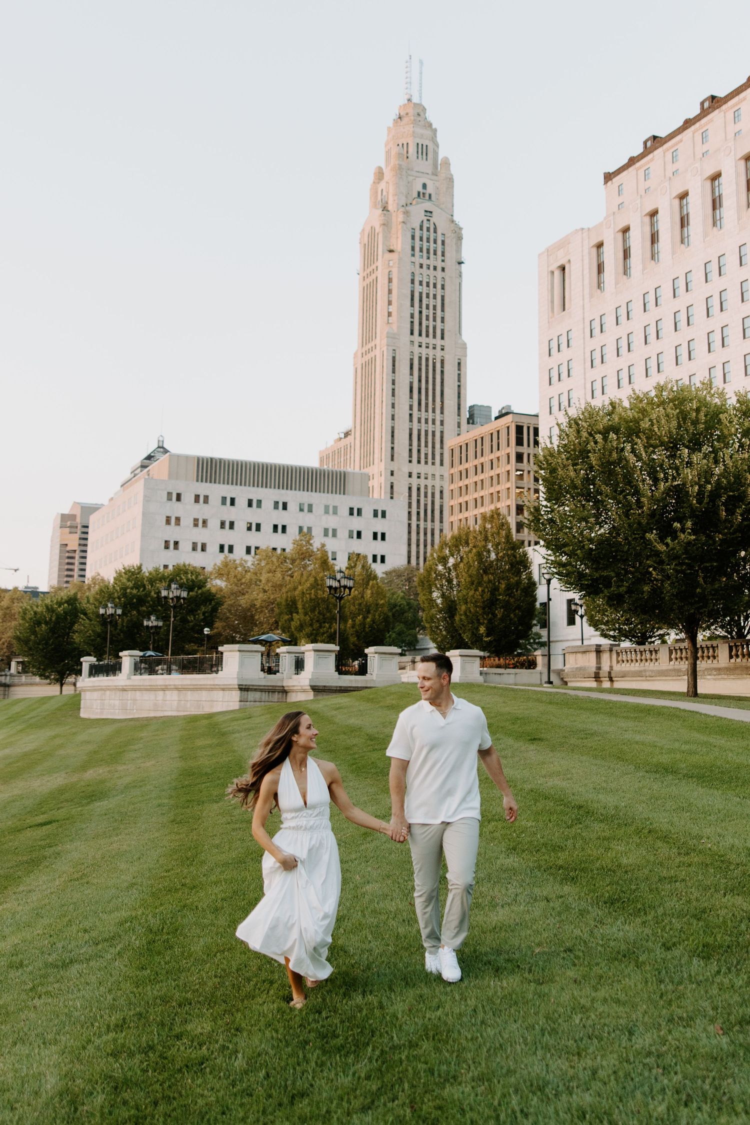 Couple holding hands and walking on green lawn with city skyline and skyscraper in the background.