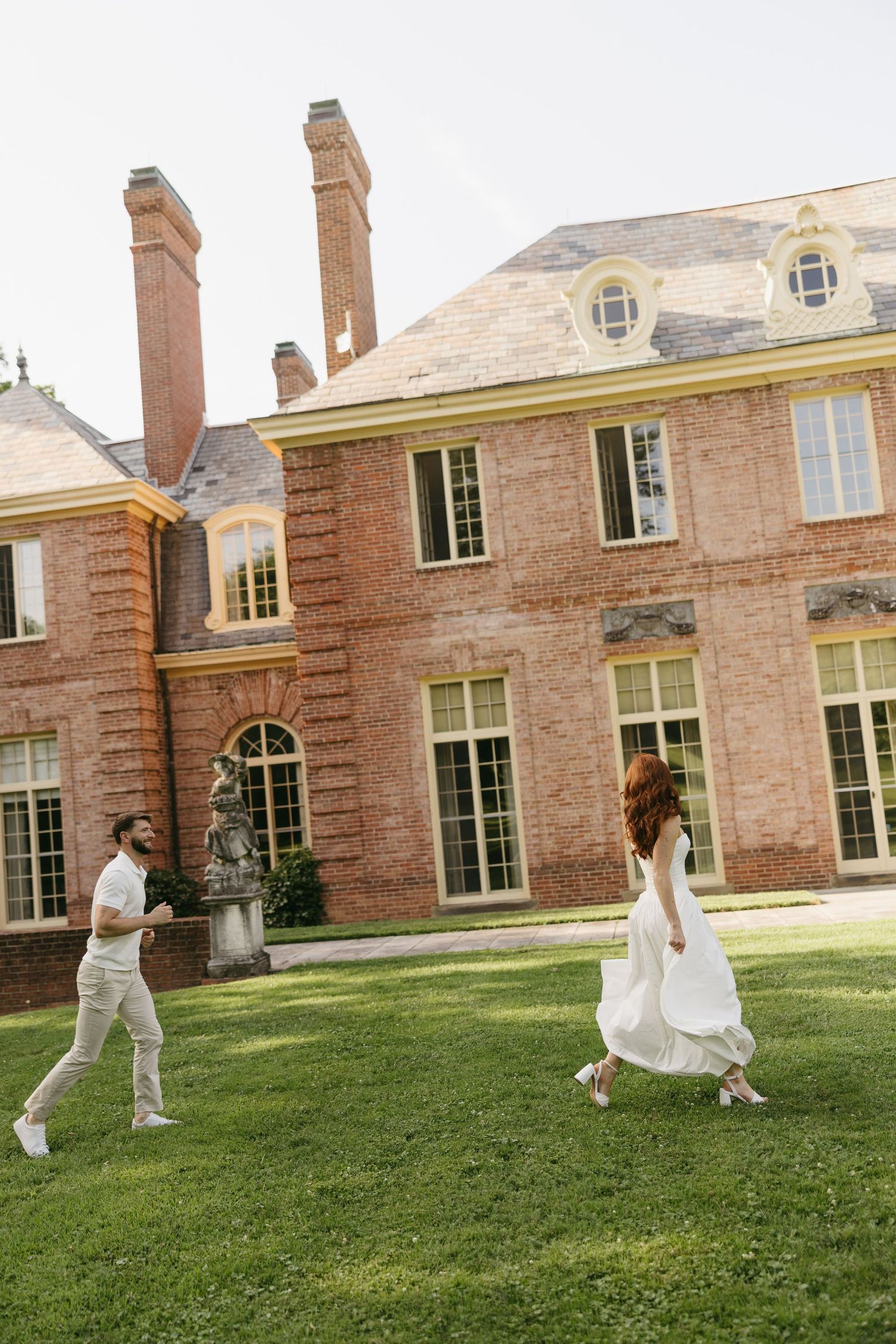 Bride in flowing white dress and groom in beige outfit run playfully on green lawn before a grand brick mansion.