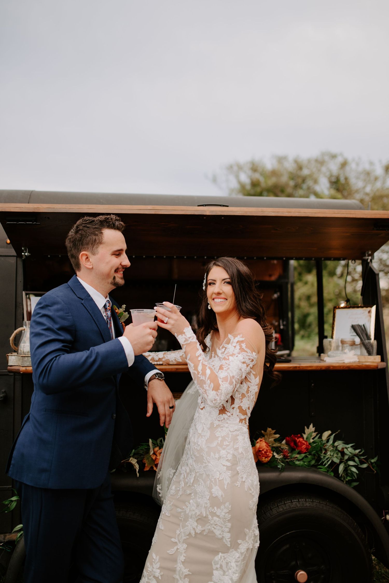 Bride and groom smiling and toasting drinks in front of a vintage black bar truck at their wedding.