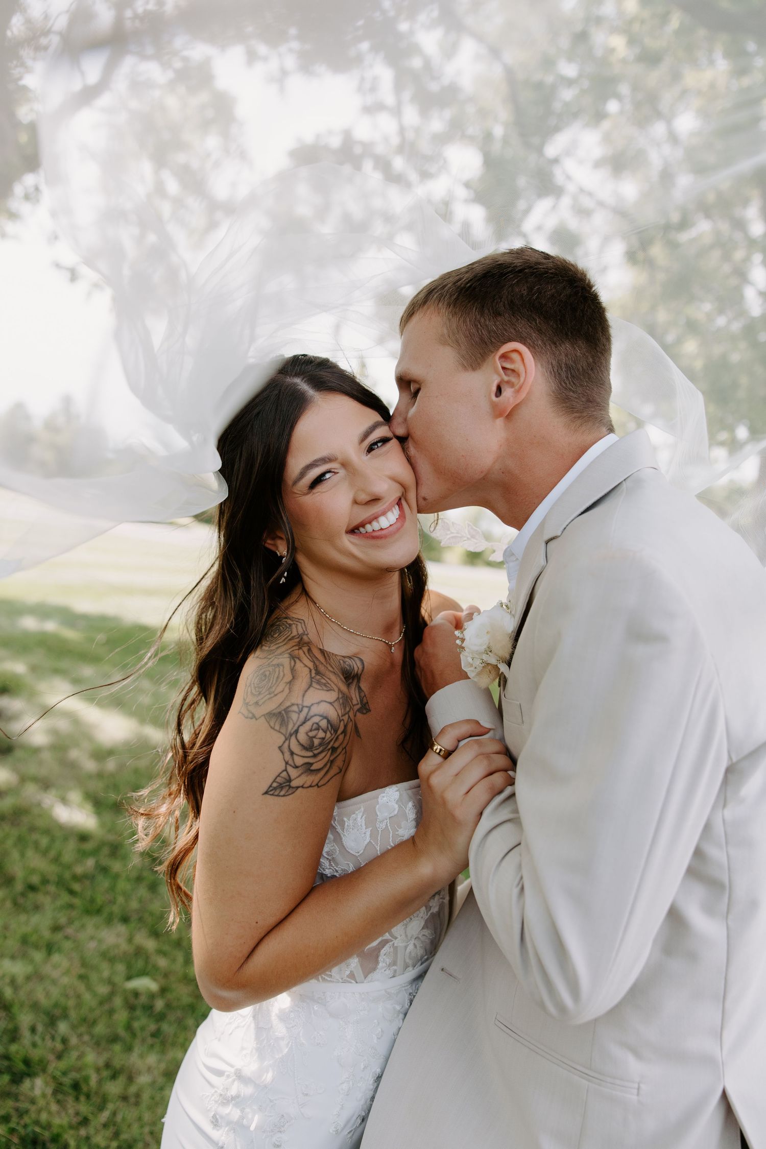 Groom kisses smiling tattooed bride on cheek as her white veil wraps softly around them.