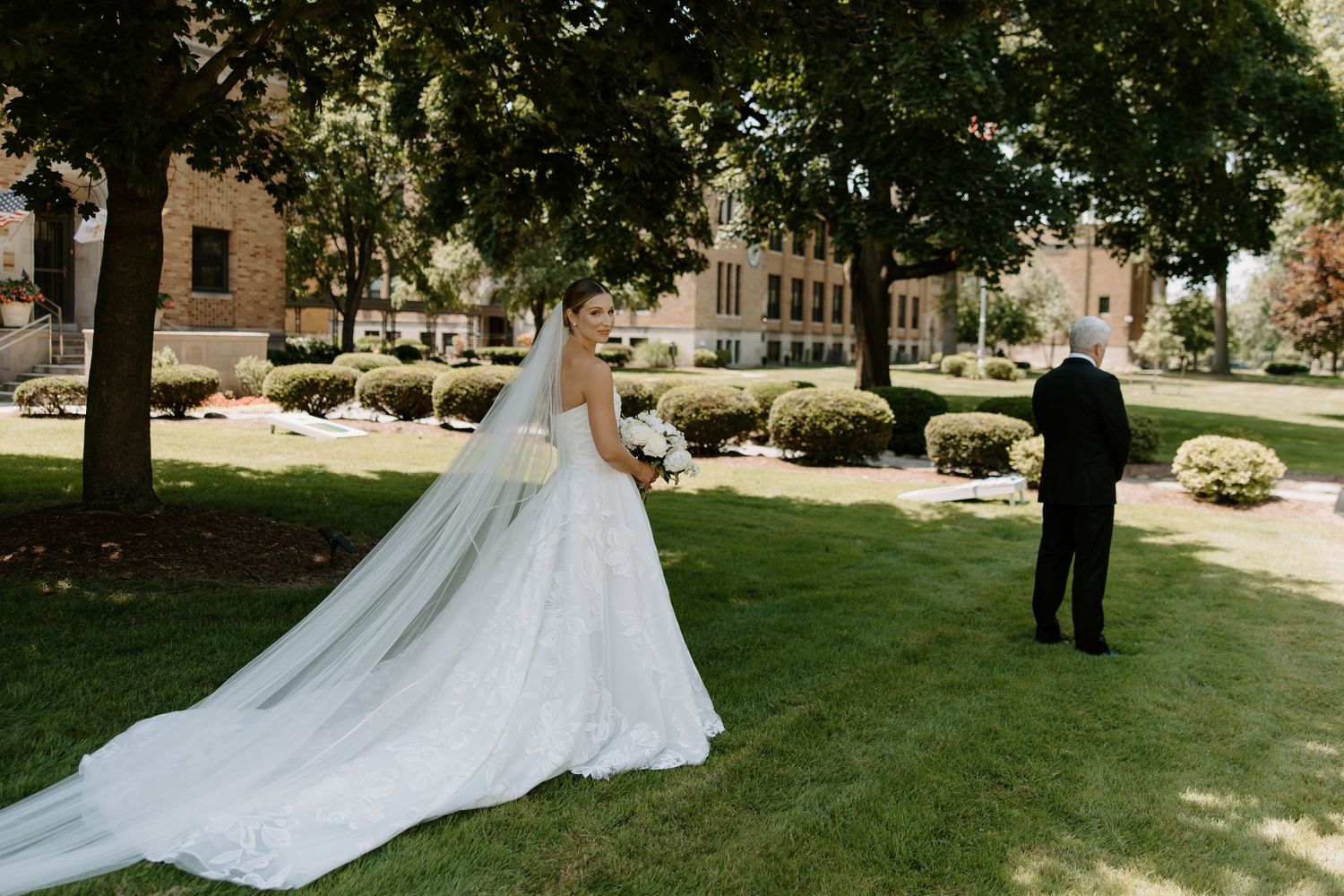 Bride in a lace wedding gown with long veil stands outdoors for a first look moment on a sunny day.