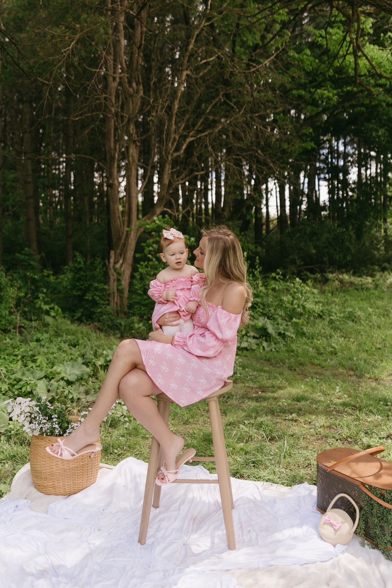 Mother in pink dress holding baby girl outdoors on white blanket near forest during picnic.