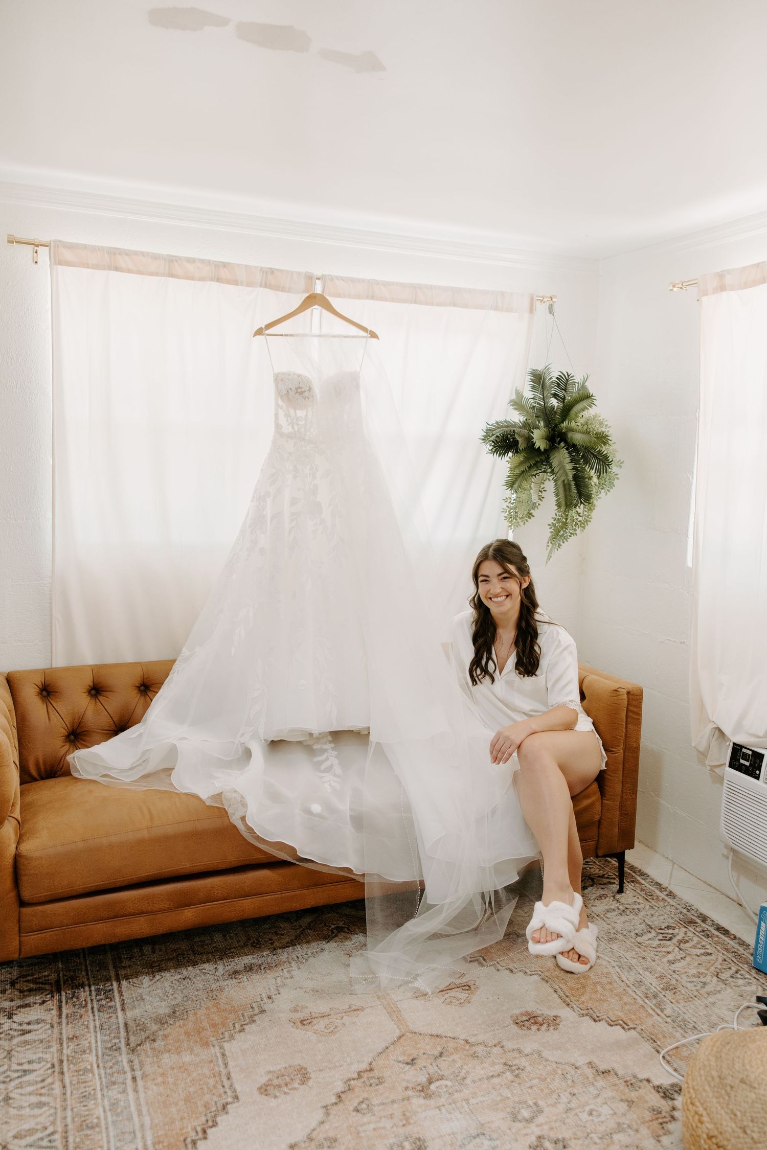 Smiling bride in white robe sitting on tan sofa beside elegant lace wedding gown hanging on wooden hanger.