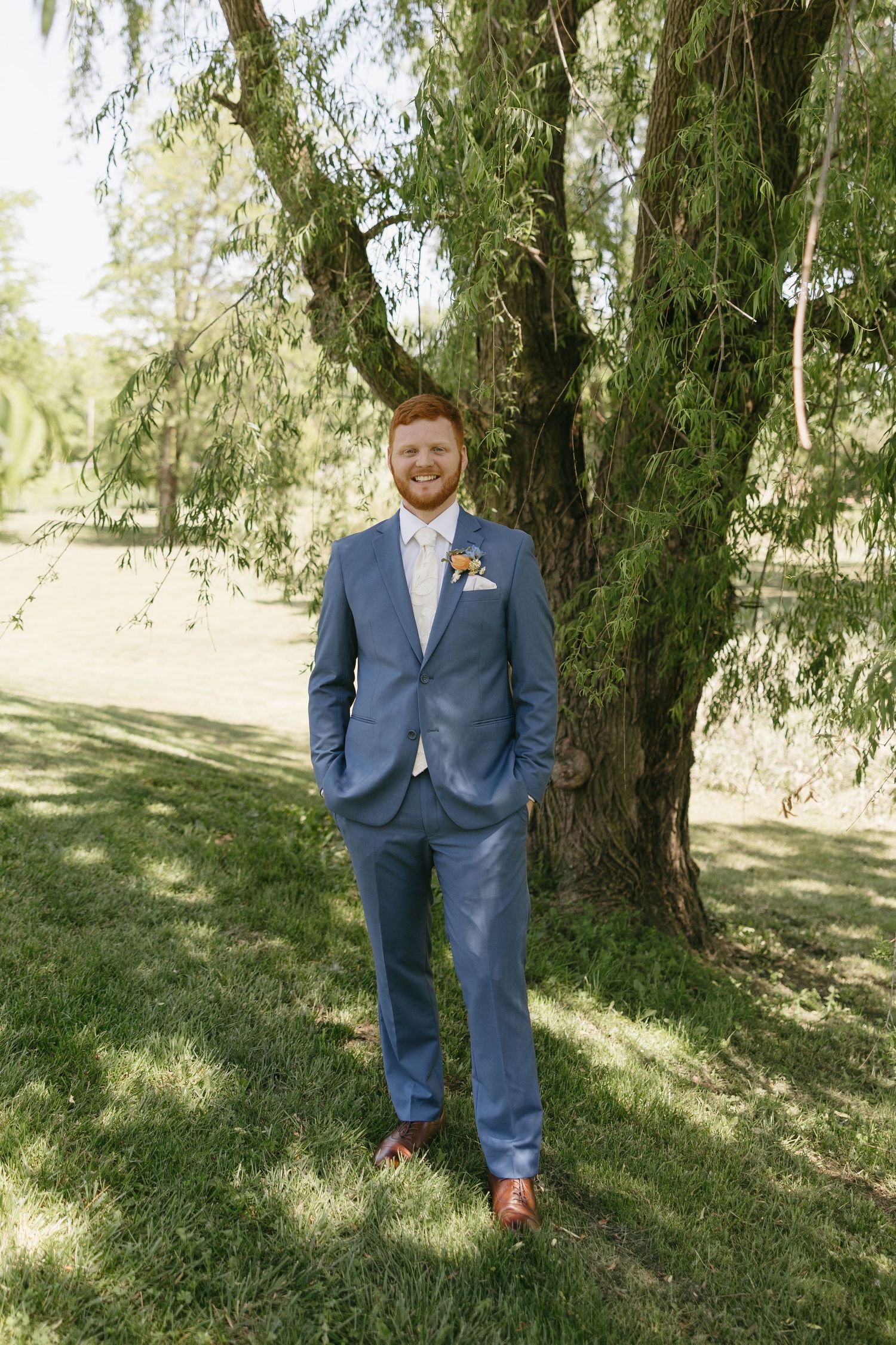 Smiling groom in blue suit with boutonniere stands under weeping willow tree on sunny wedding day.