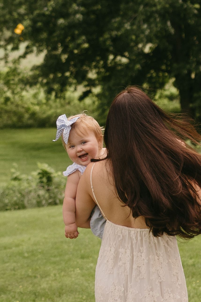 Mother with long brown hair holds laughing baby girl with white bow in sunny green backyard.