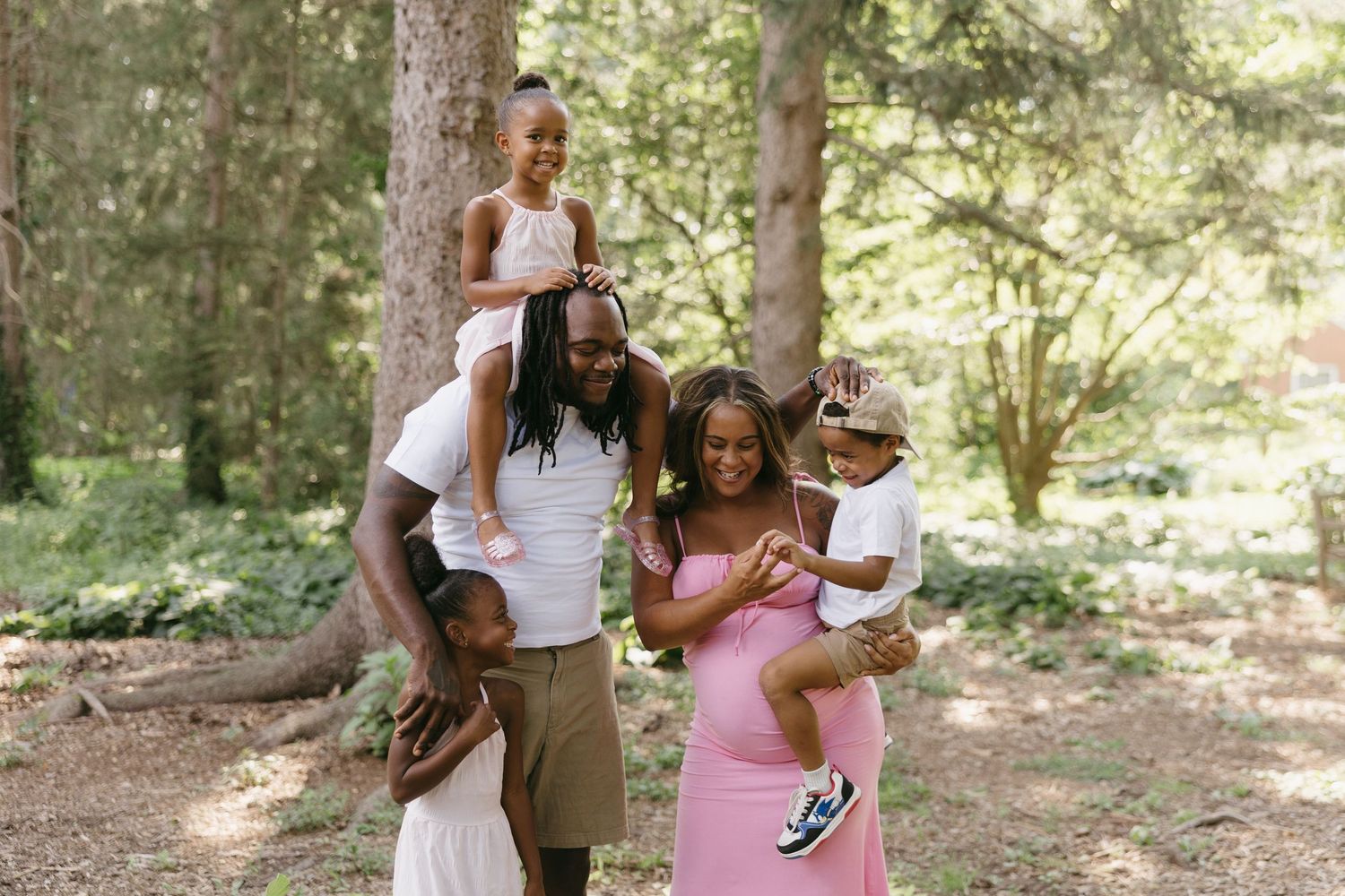 Happy Black family of five laughs together in lush green forest, parents holding young children.
