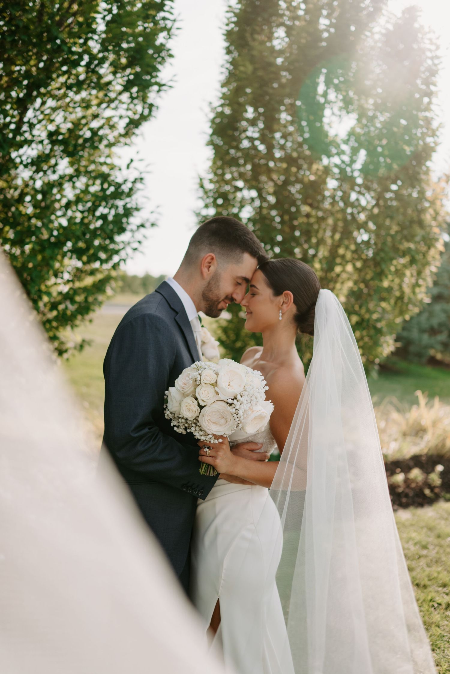 Bride and groom touching foreheads tenderly outdoors, bride holding white rose bouquet with veil flowing in sunlight.