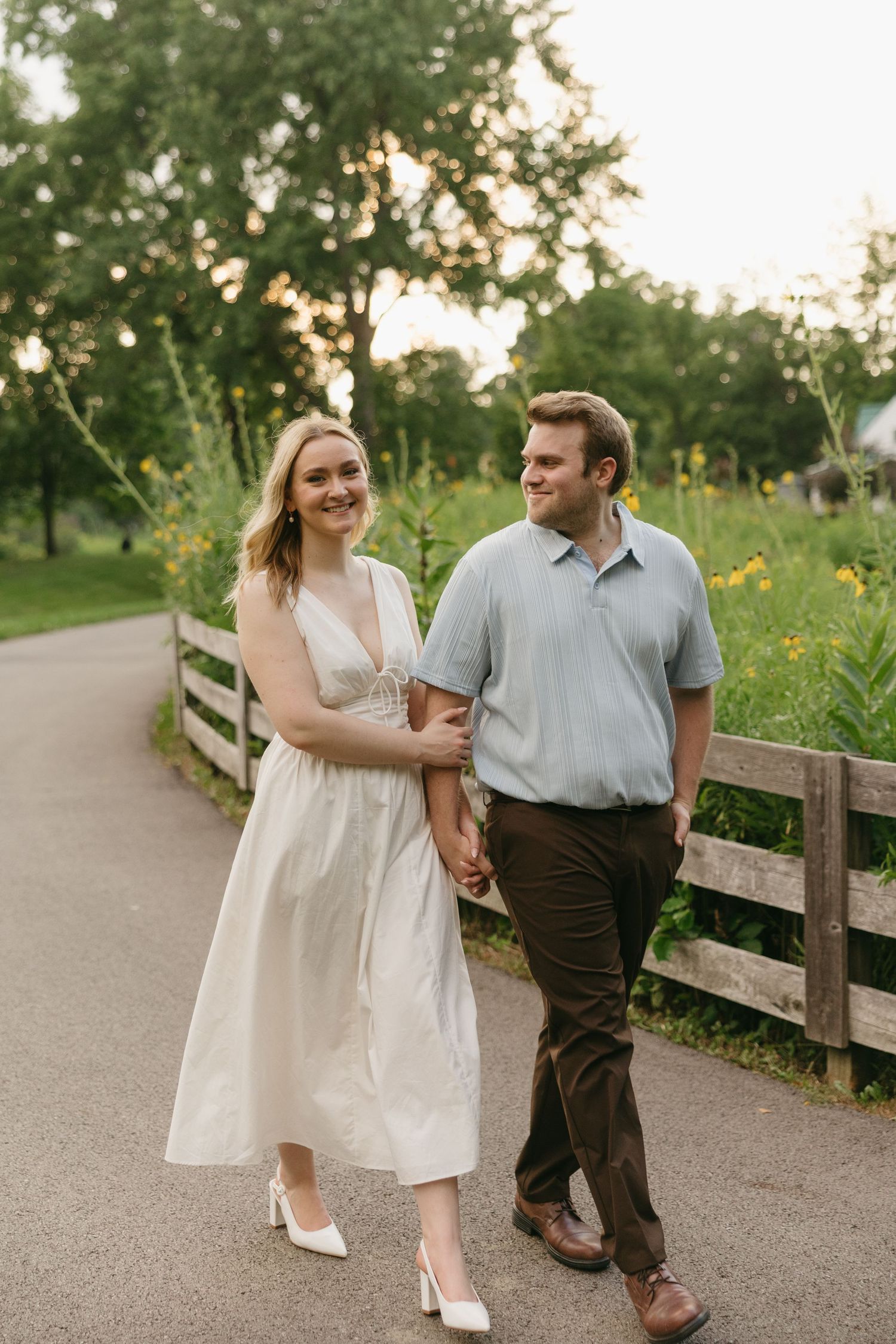 Smiling couple walking hand in hand along a path lined with wildflowers and a wooden fence.