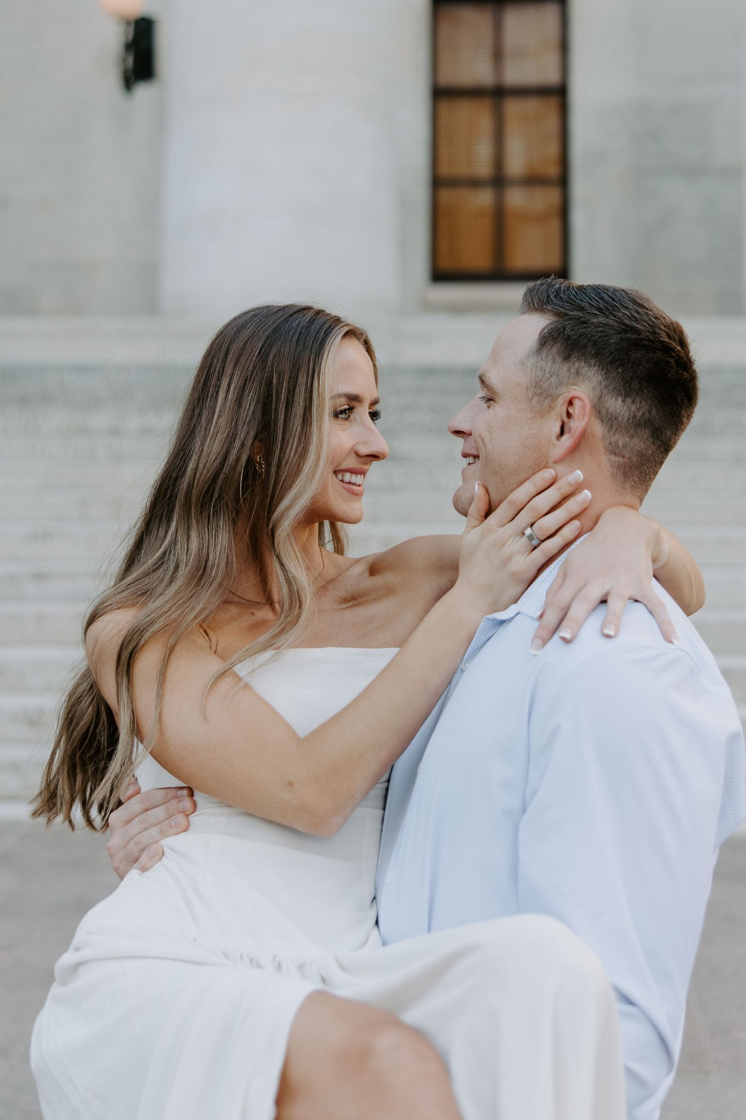 Woman in white dress cradling mans face while being held, both smiling at each other near marble columns.