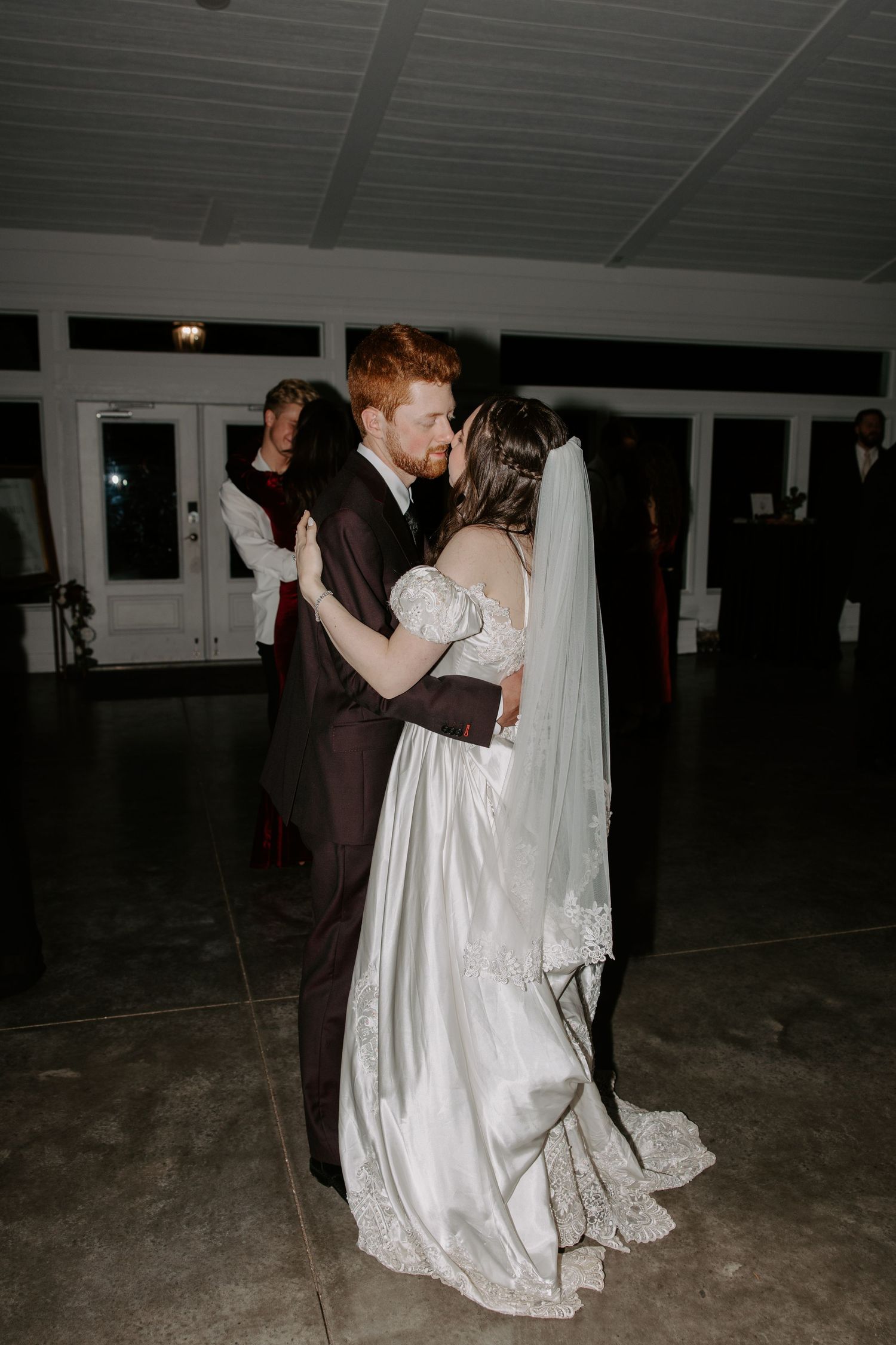 Red-haired groom in dark suit shares first dance kiss with veiled bride in white satin gown at reception.