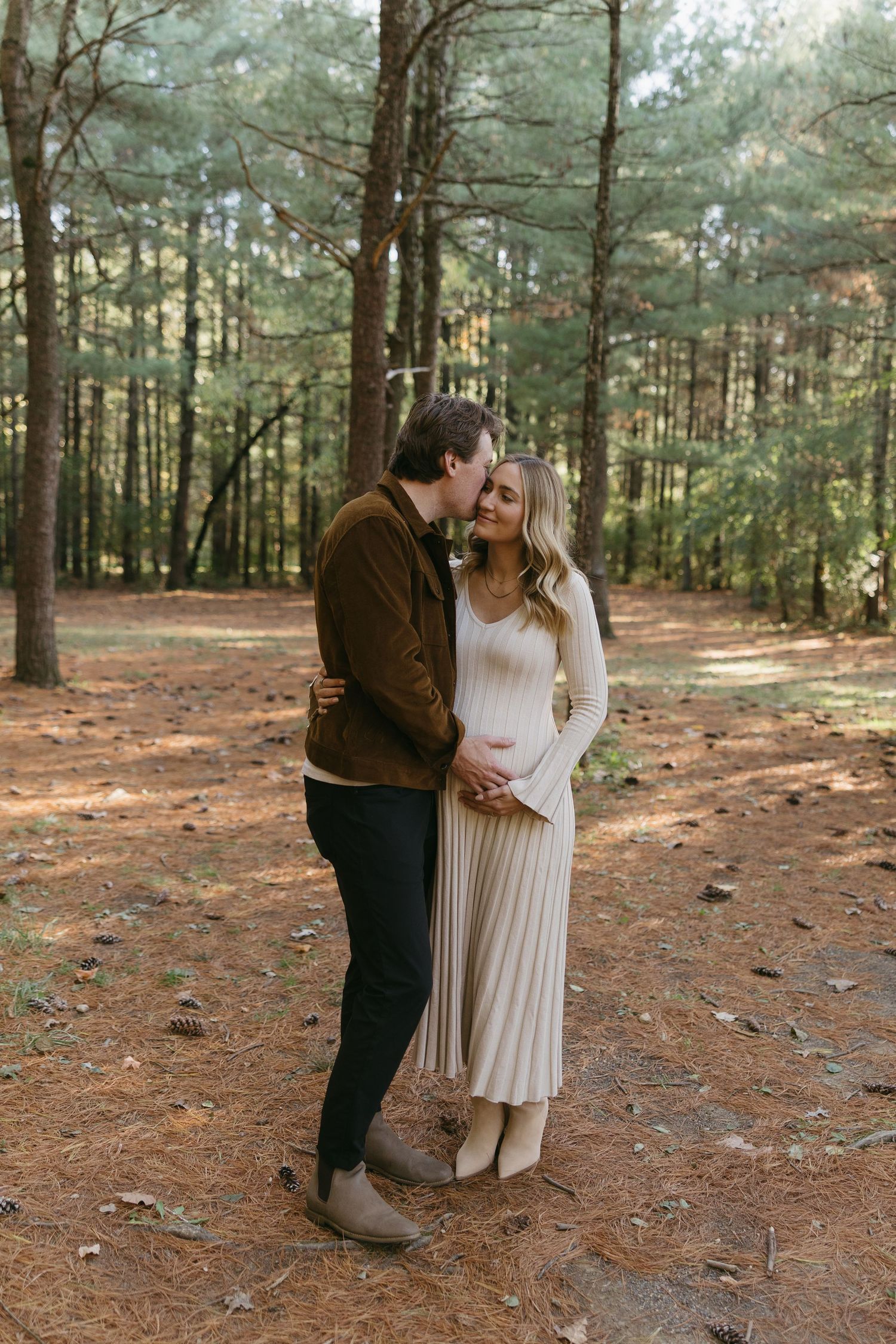 Couple sharing a tender moment in a pine forest, woman in cream pleated dress cradling baby bump during maternity photoshoot.