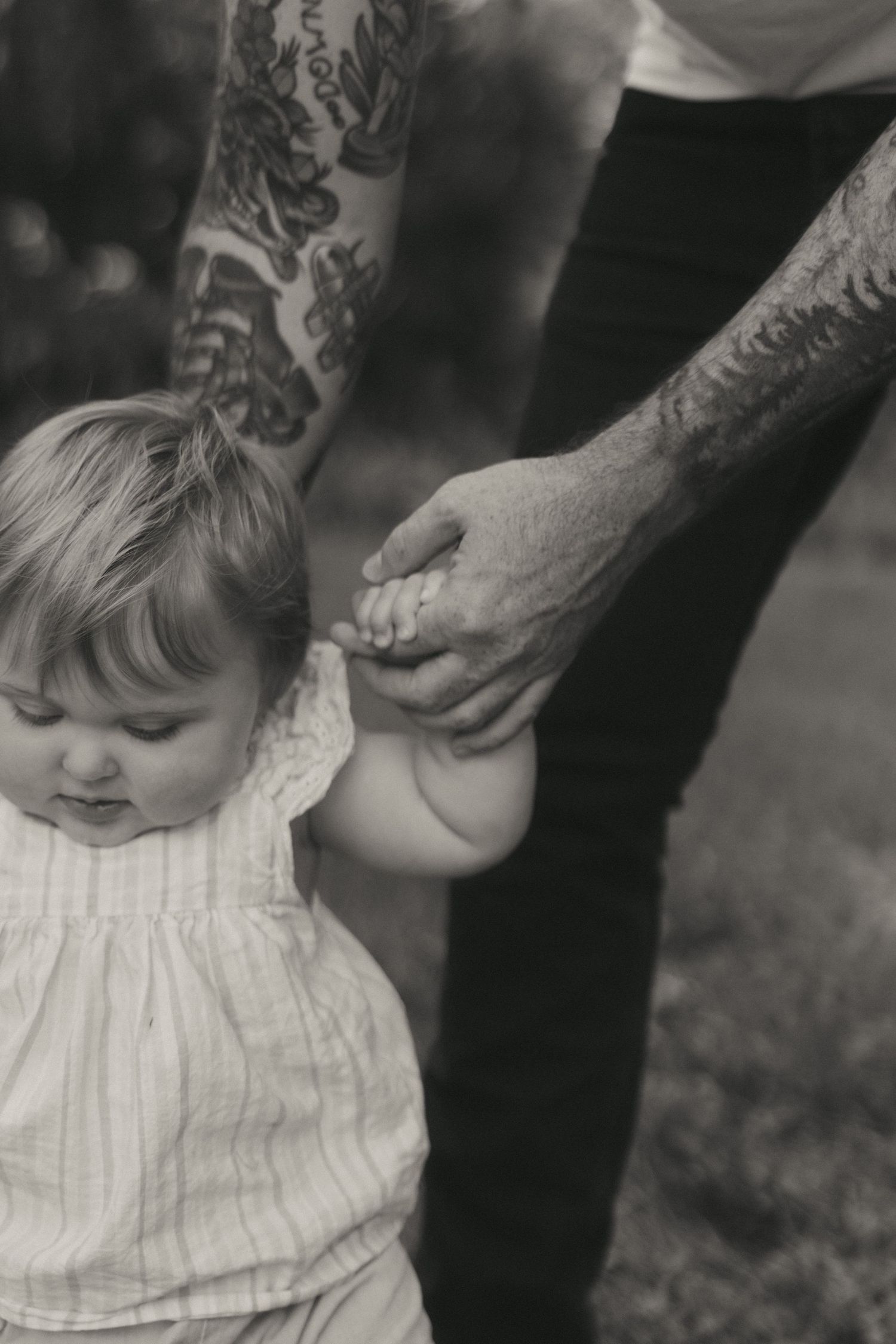 Black and white photo of tattooed father holding baby girls hands as she learns to walk outdoors.