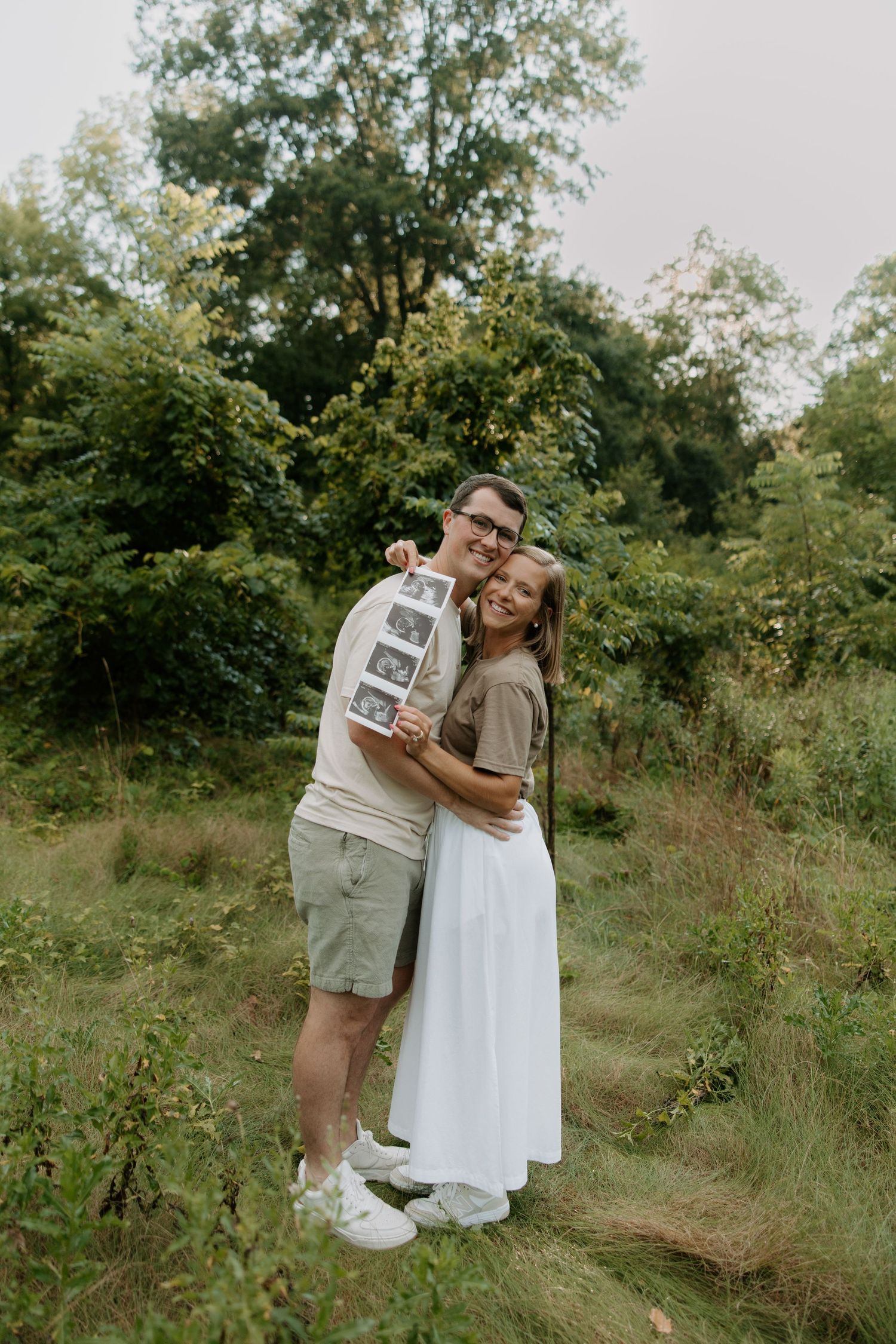 Happy couple hugging outdoors in a green meadow while holding ultrasound photos for a pregnancy announcement.
