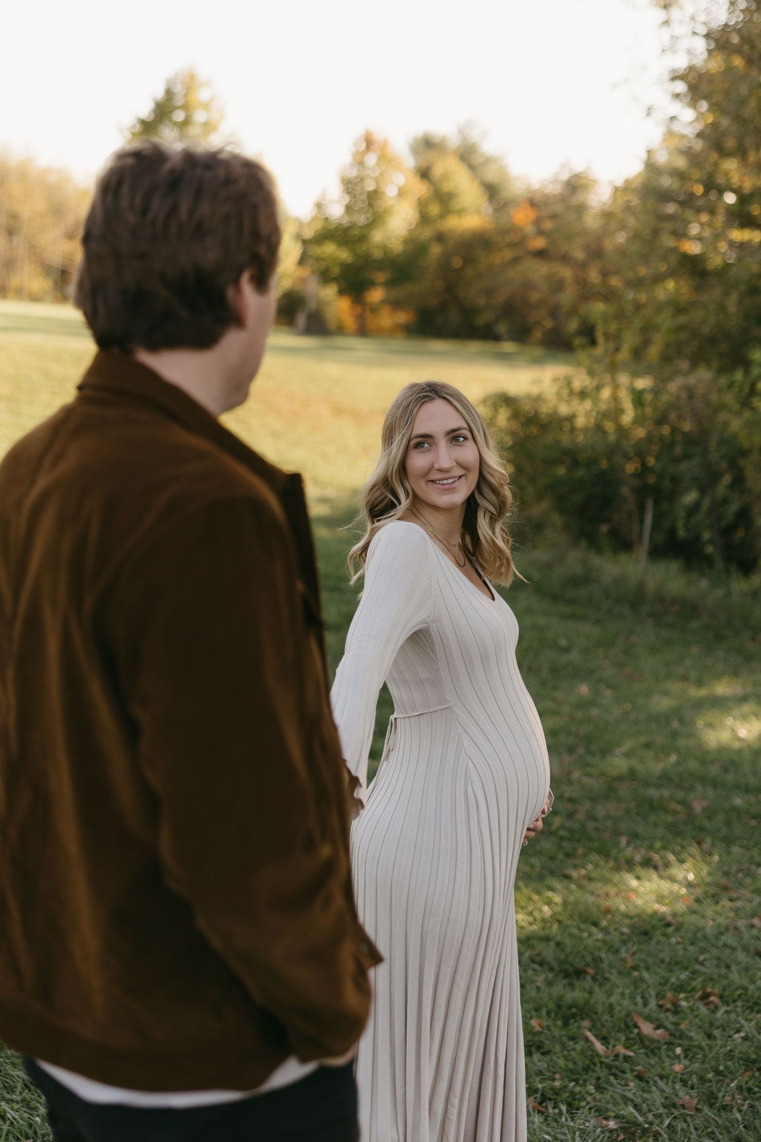 Pregnant woman in cream ribbed dress smiles over shoulder at partner during golden hour maternity photoshoot outdoors.