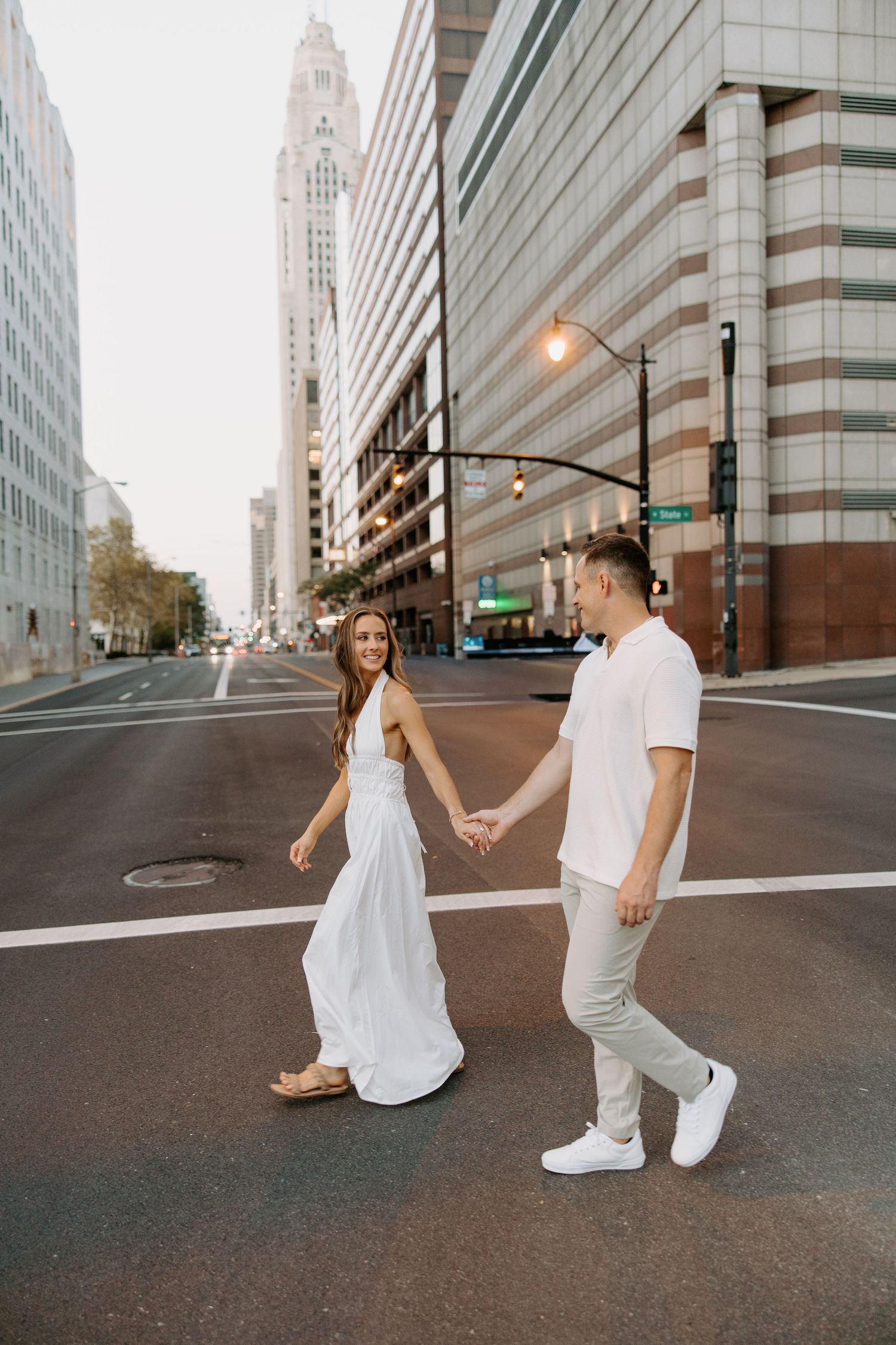 Couple holding hands crossing an urban street with tall city buildings and a skyscraper in the background.