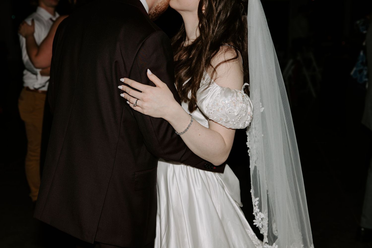 Bride and groom share intimate kiss on dark dance floor, bride wearing white gown and veil with ring visible.
