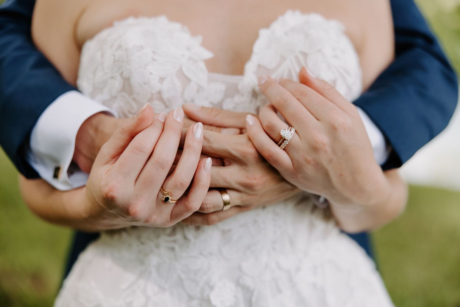 Close-up of bride and groom hands showing wedding rings, bride in lace dress, groom in blue suit.