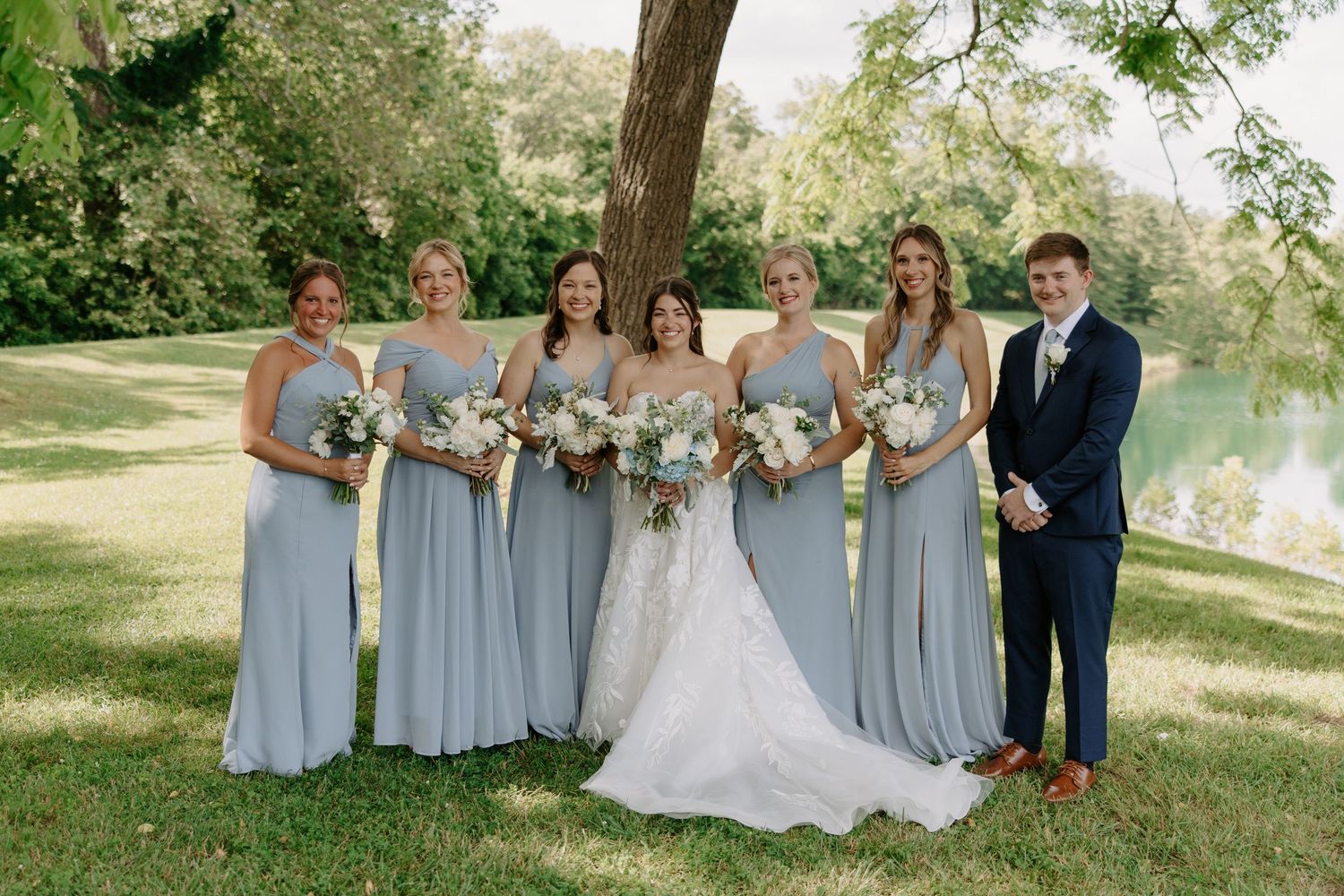 Bride in white lace gown posing with bridesmaids in dusty blue dresses and groomsman in navy suit outdoors.