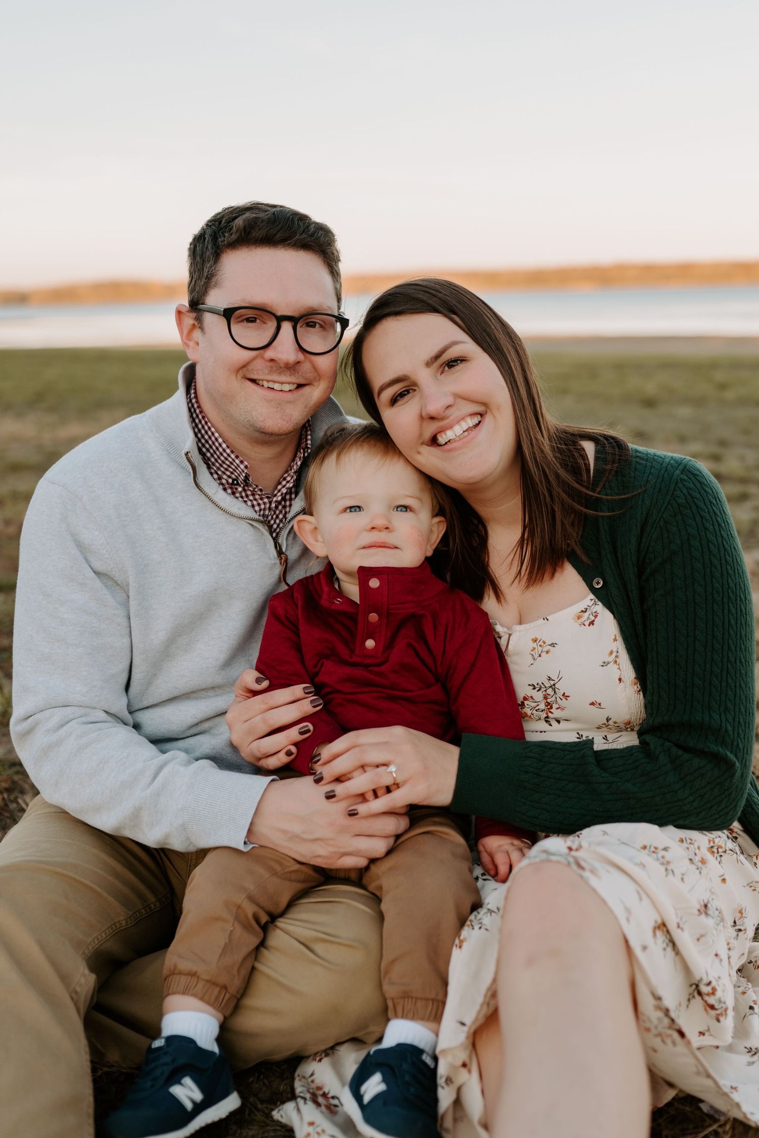 Happy family portrait with parents and toddler sitting outdoors near a lake at sunset.