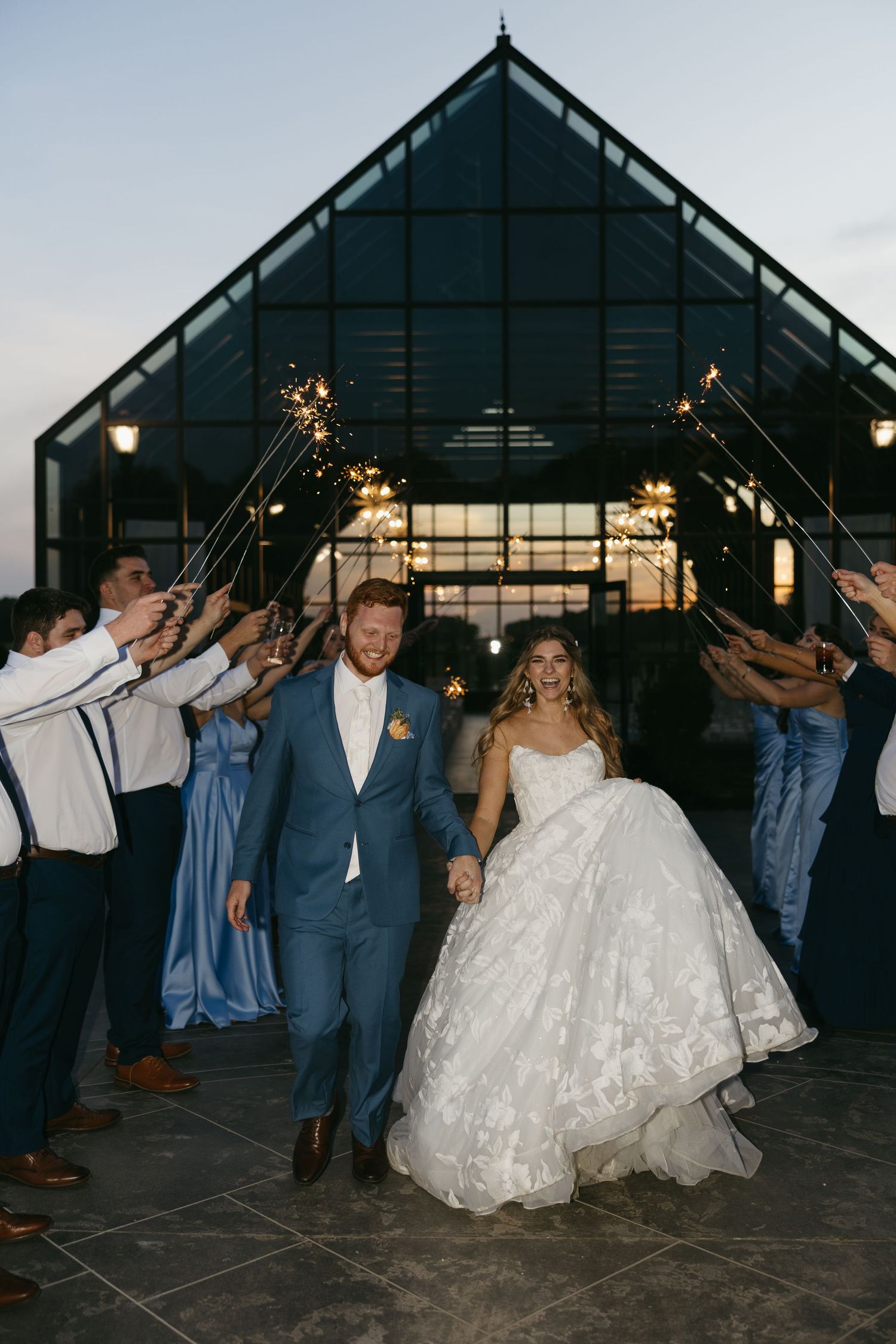 Newlyweds exit greenhouse venue through sparkler tunnel at dusk, groom in blue suit.
