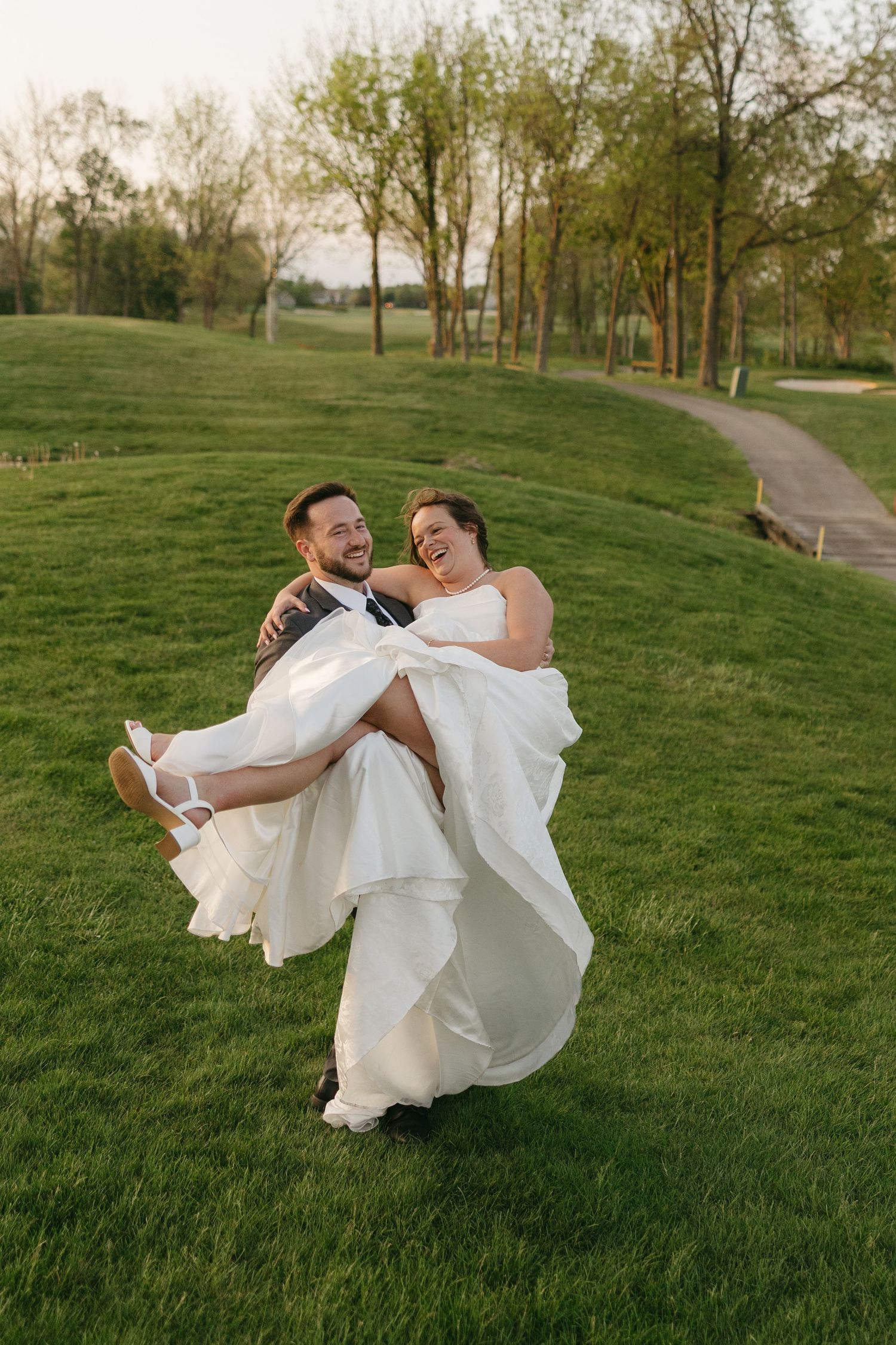 Laughing groom carries his joyful bride in a white gown across a lush green golf course.