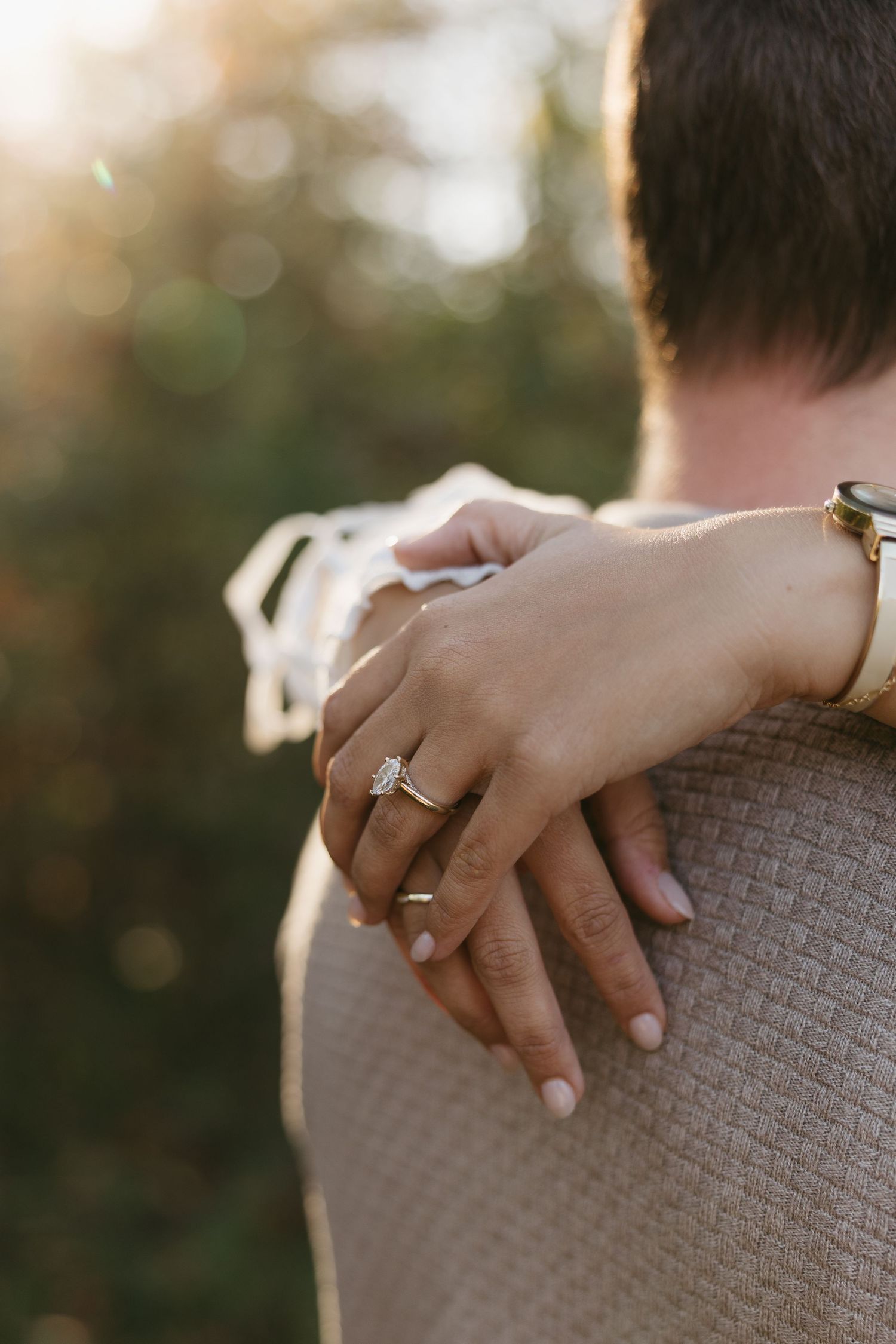Close-up of a womans hand with a diamond engagement ring resting on a mans shoulder outdoors.