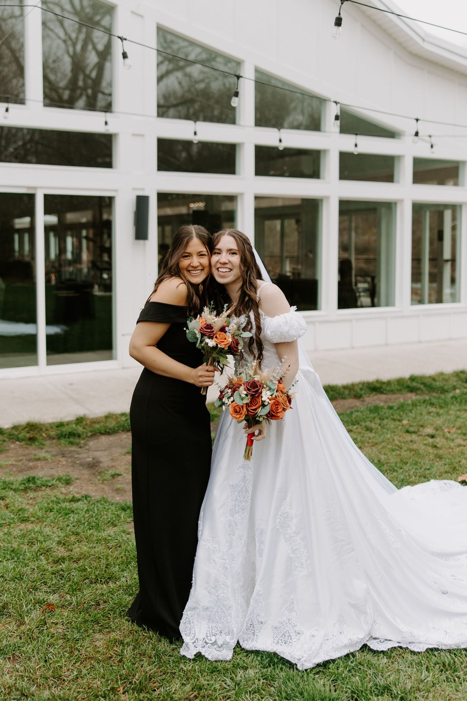 Bride in white lace gown holding bouquet poses with bridesmaid in black off-shoulder dress outside white venue.