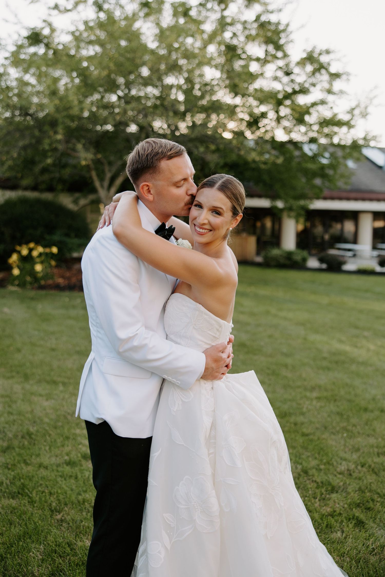 Bride and groom embracing on green lawn, groom in white tuxedo kissing bride's cheek as she smiles in strapless gown.