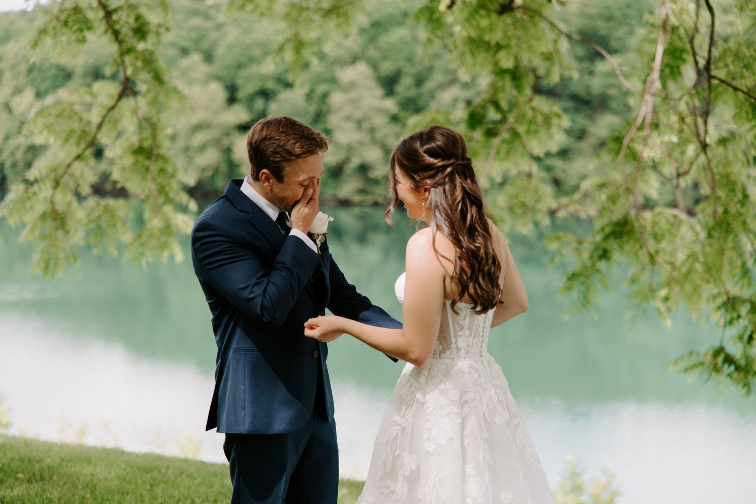Groom wiping tears holding bride's hands in lace gown by lakeside, surrounded by lush green trees.