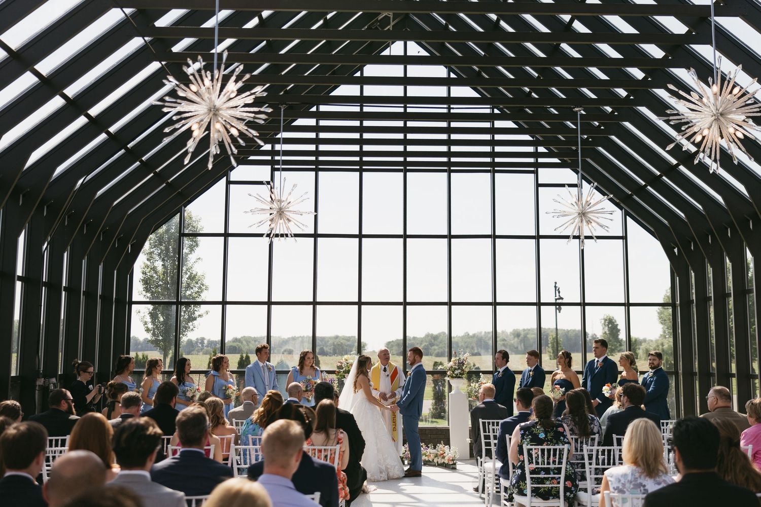 Elegant wedding ceremony in a glass greenhouse venue with star chandeliers and scenic views.