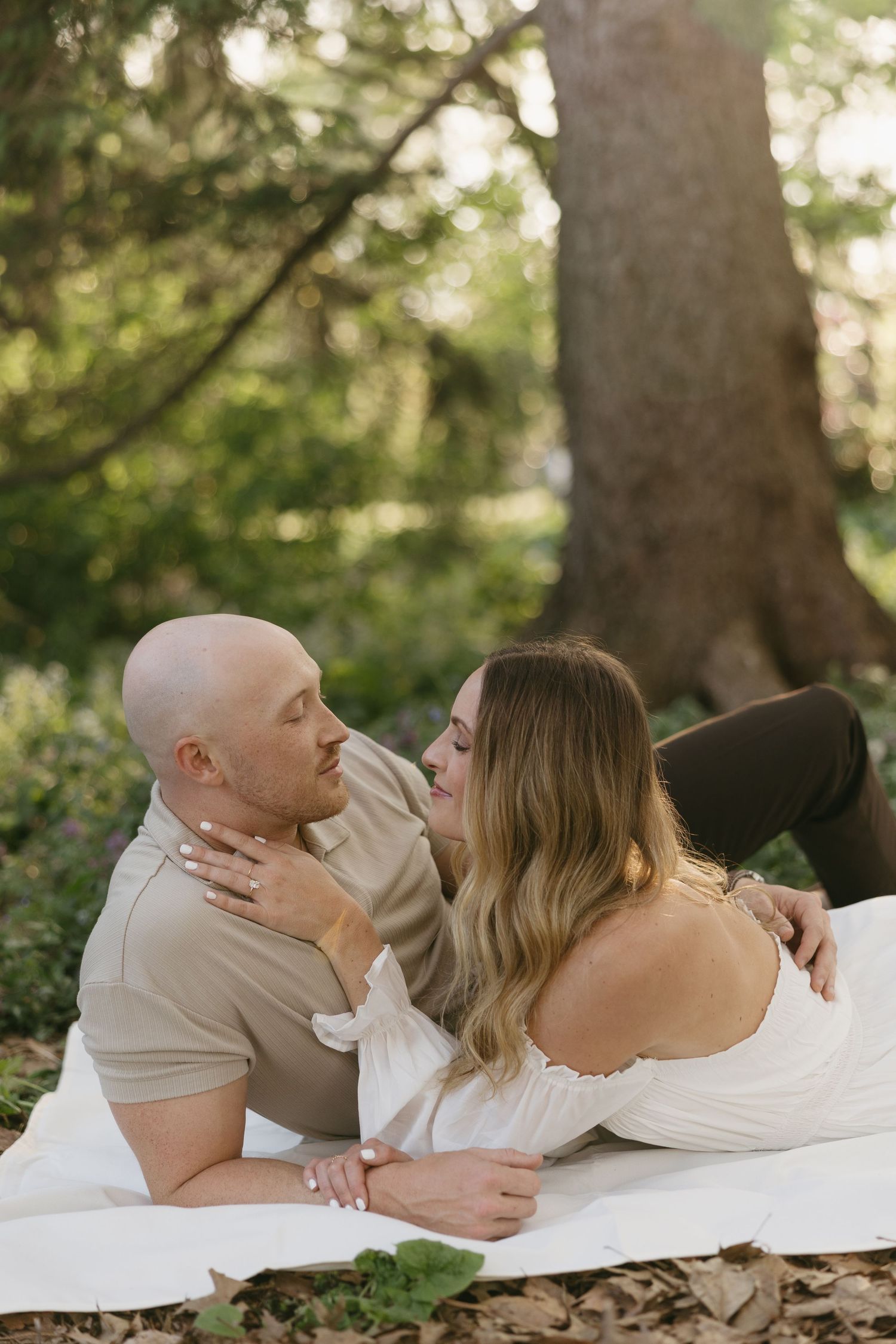 Couple lying on a white blanket in a forest, gazing at each other with a warm golden light.