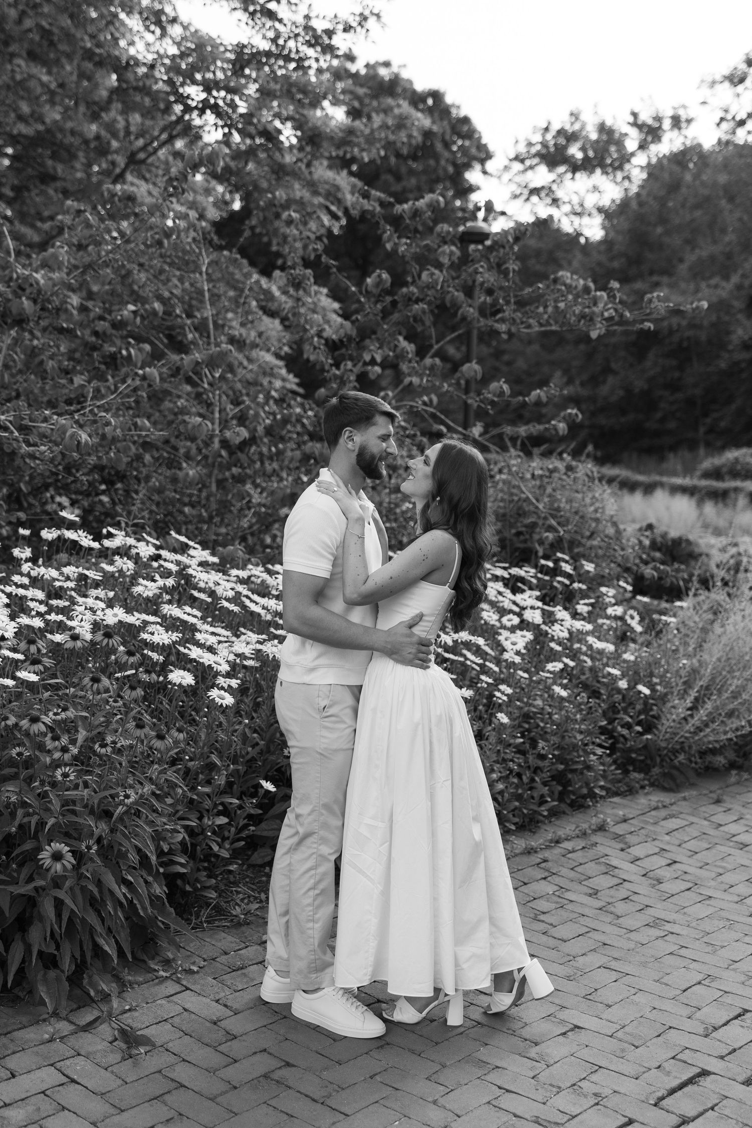 Black and white photo of couple embracing near wildflowers in a lush garden setting.
