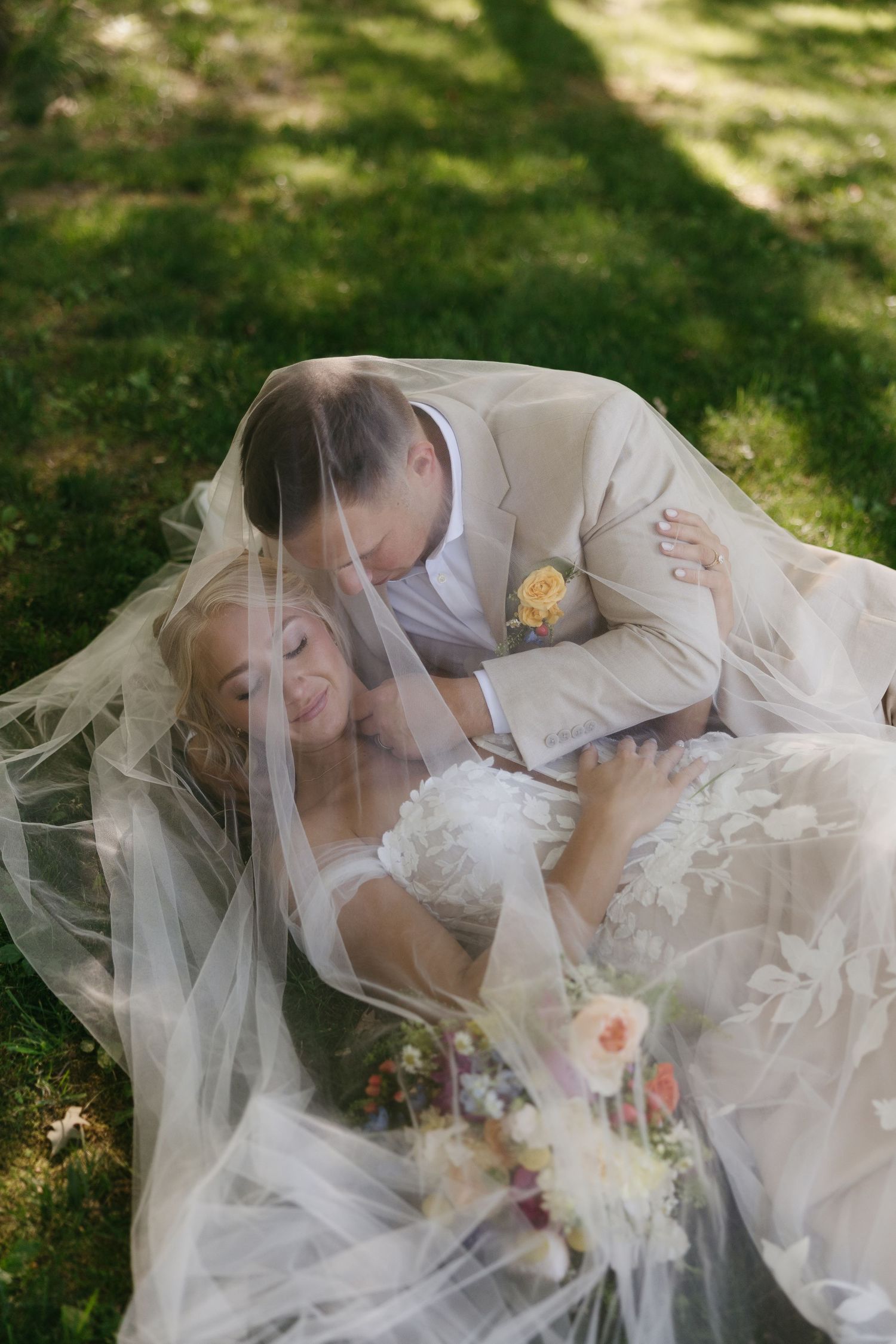 Bride and groom lying on grass under wedding veil, sharing a romantic moment outdoors.