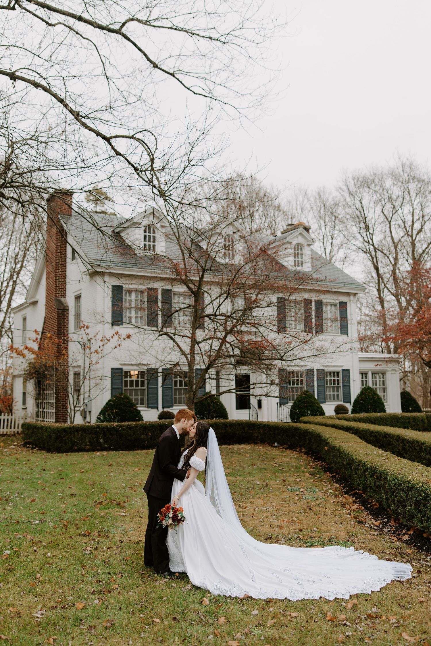 Bride and groom kissing in front of elegant white colonial manor surrounded by manicured hedges in autumn.