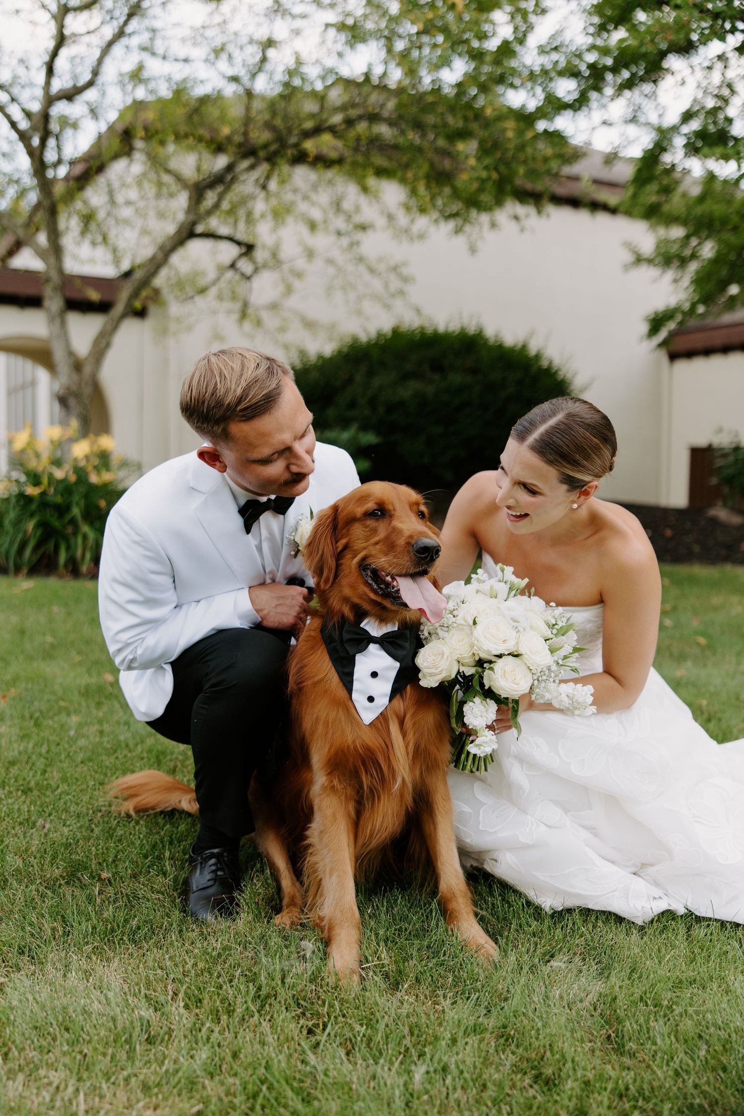 Newlyweds smile while petting their golden retriever dressed in a tuxedo bib on their wedding day.