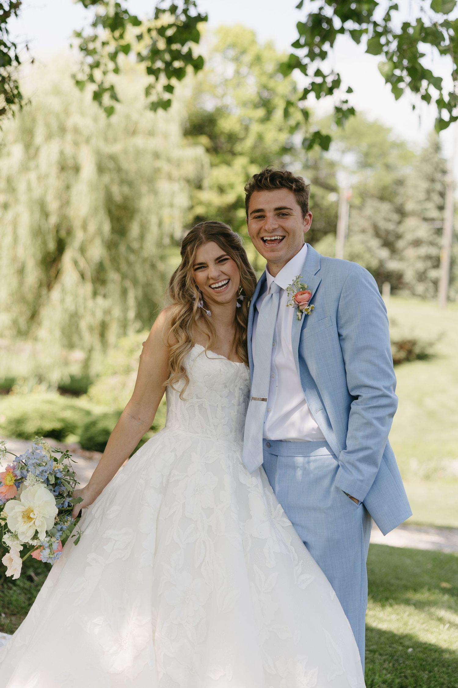 Happy couple posing outdoors, bride in white gown holding bouquet, groom in light blue suit with floral boutonniere.