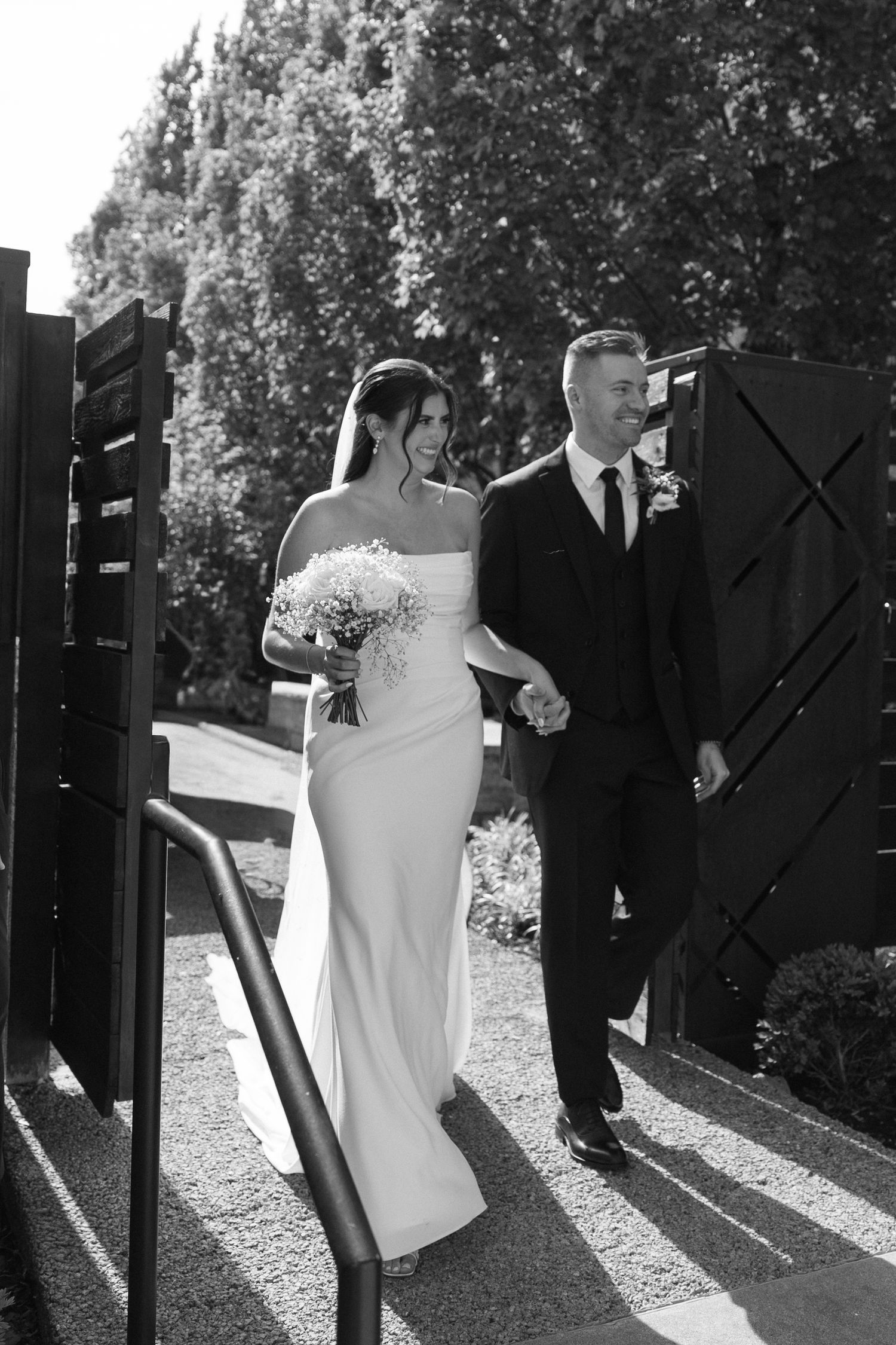 Black and white photo of bride and groom walking hand in hand through elegant venue gates.