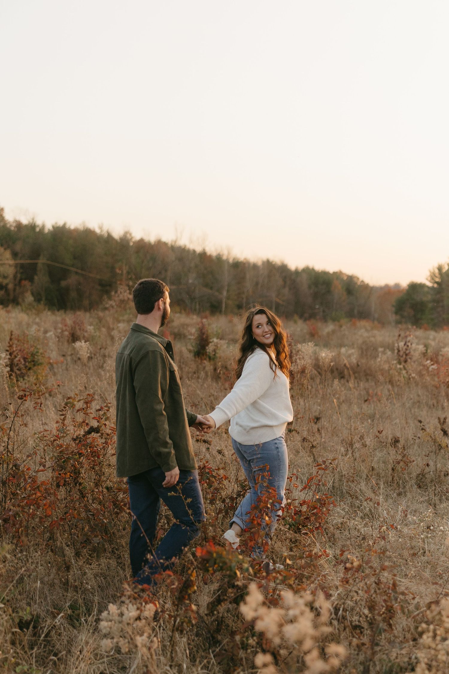 Couple holding hands walking through an autumn field at golden hour, woman in white sweater smiling back at camera.