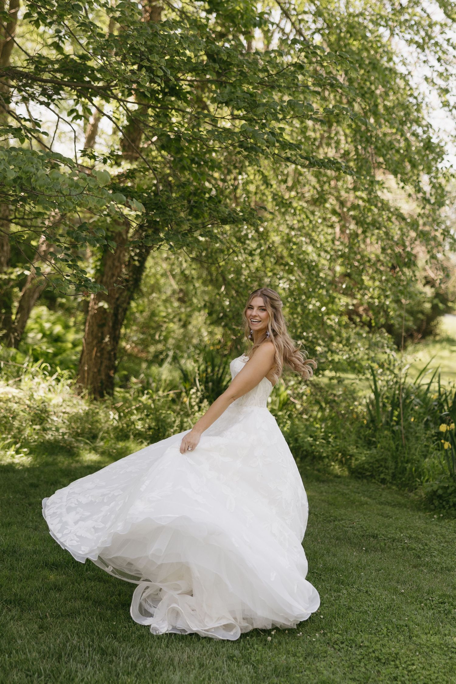 Bride in a flowing white tulle wedding gown twirling outdoors in a lush green garden setting.